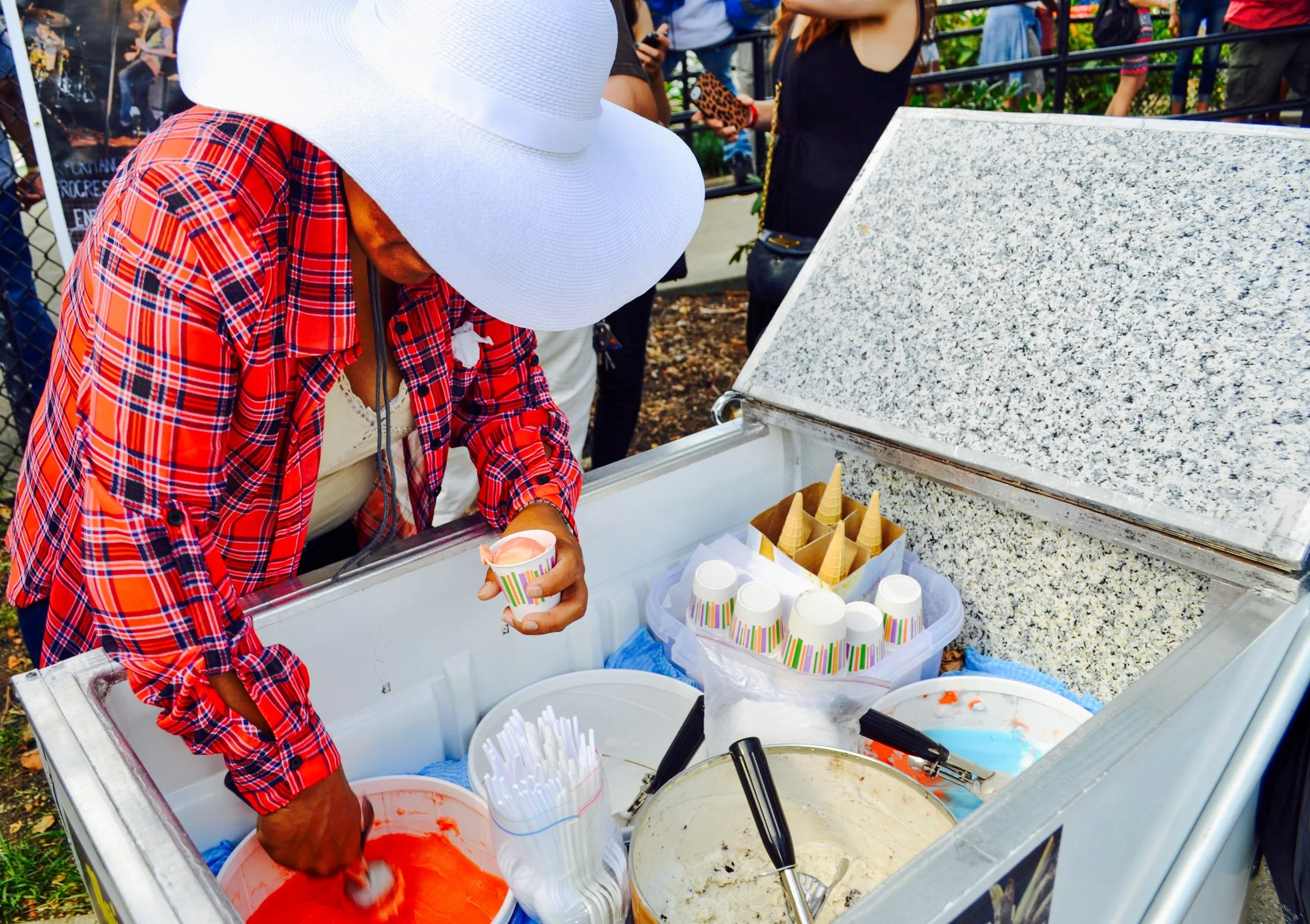 A person wearing a large white sun hat and a red plaid shirt scoops ice cream from a freezer at an outdoor event. There are ice cream cones and cups, with several containers of ice cream in the freezer. Other people are gathered around in the backgro