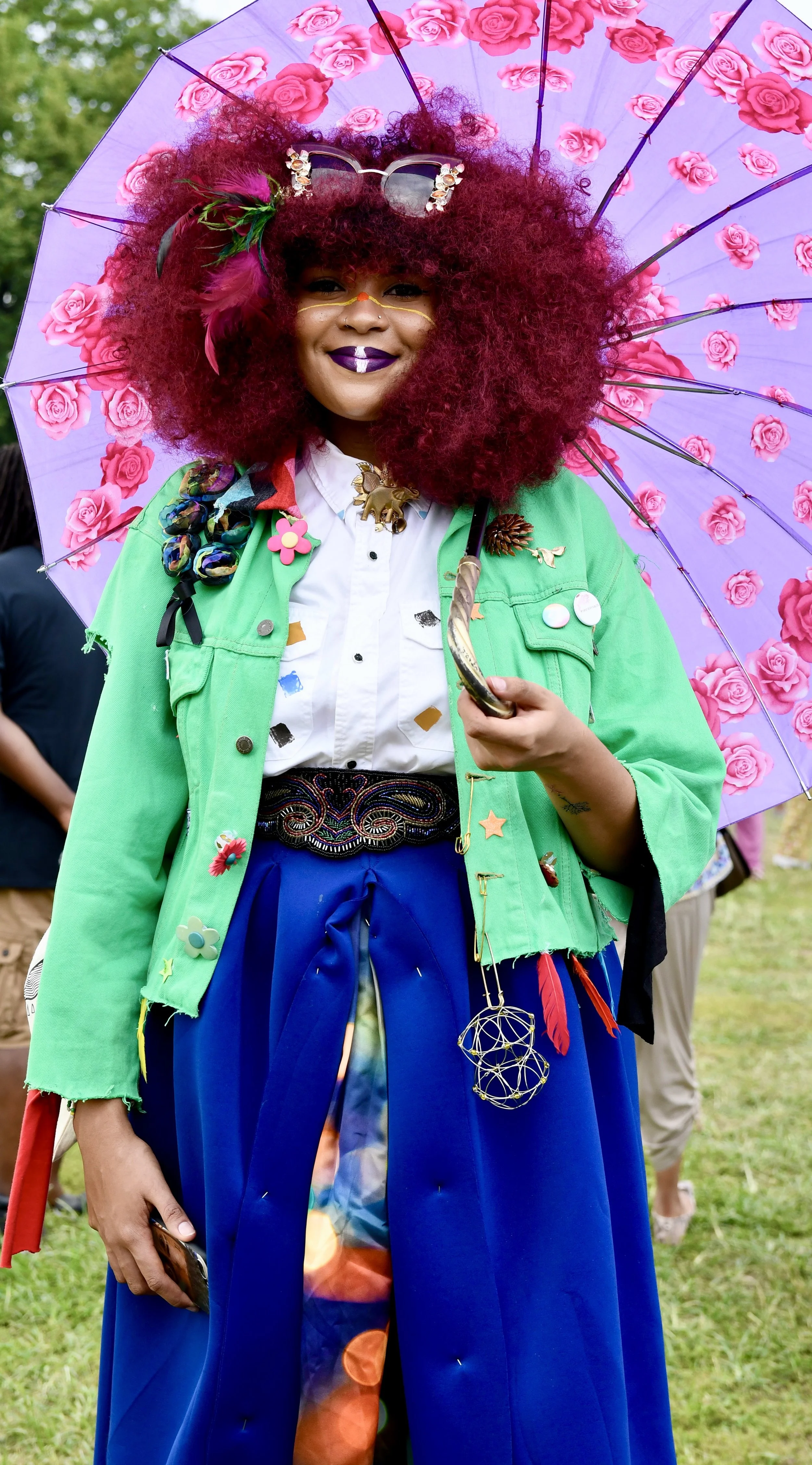 A woman with curly vibrant red hair wearing colorful eclectic accessories, glasses on her head, smiling, holding a purple umbrella with pink roses, dressed in a green jacket, and a blue skirt with a celestial pattern. AfroPunk Brooklyn 2015