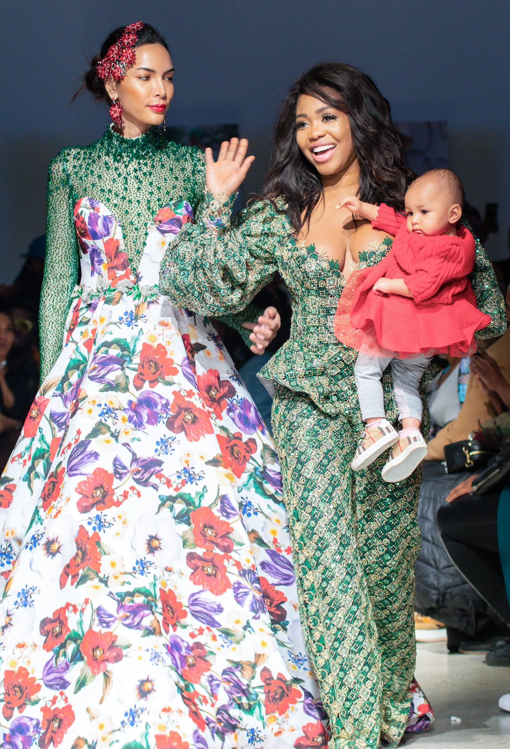 Three women, two in colorful, patterned garments and one child, are in a public setting. One woman, with long dark hair, is smiling and holding the child, who wears a red outfit. The woman next to her appears to be waving or gesturing. Designer, Ese.