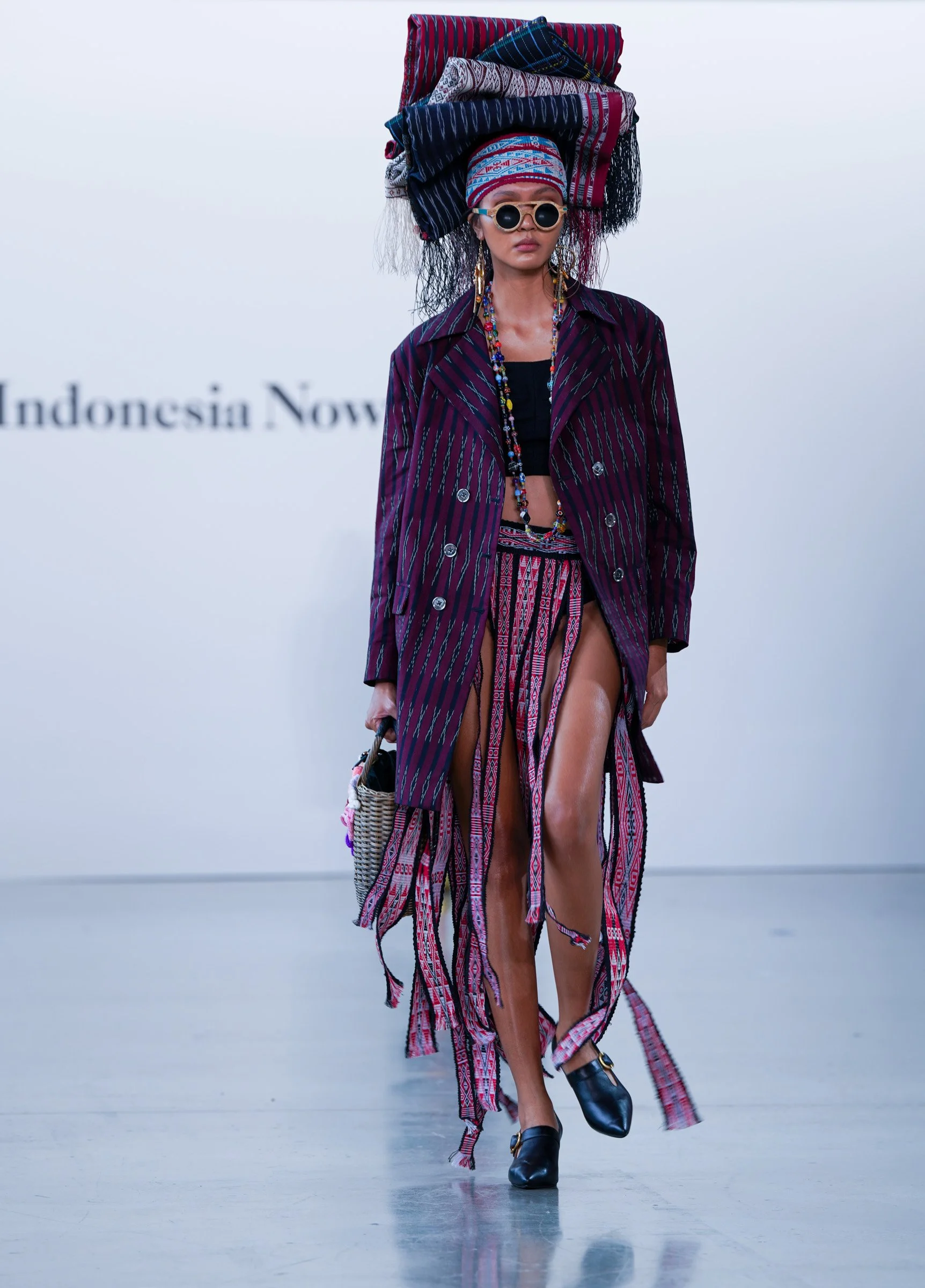 Model walking on runway wearing layered, patterned clothing with accessories and an elaborate headdress made of textiles.
