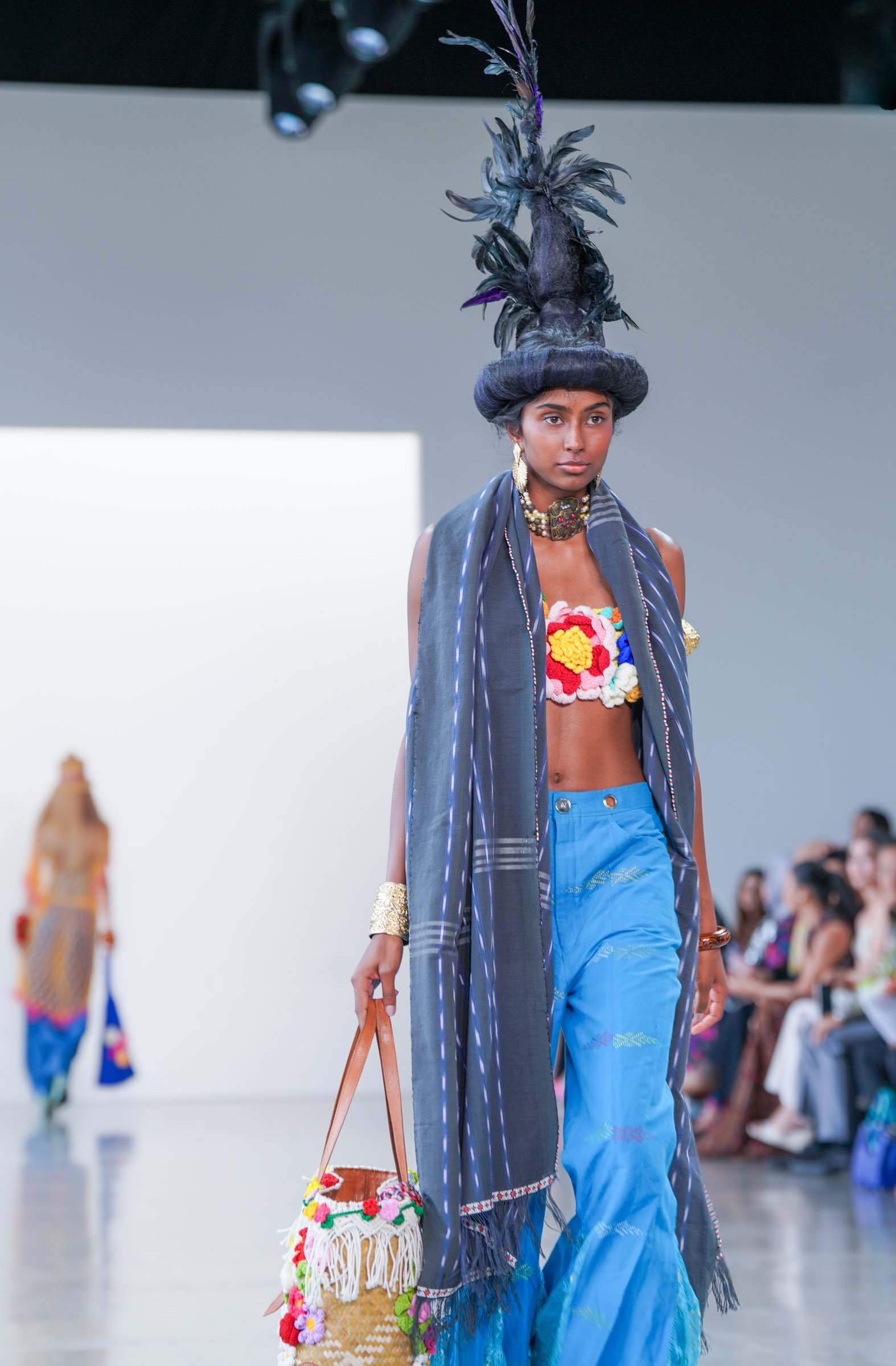 Model walking down runway at fashion show wearing a colorful, floral-embellished top, blue pants, and a large feathered hat, carrying a woven bag decorated with flowers.