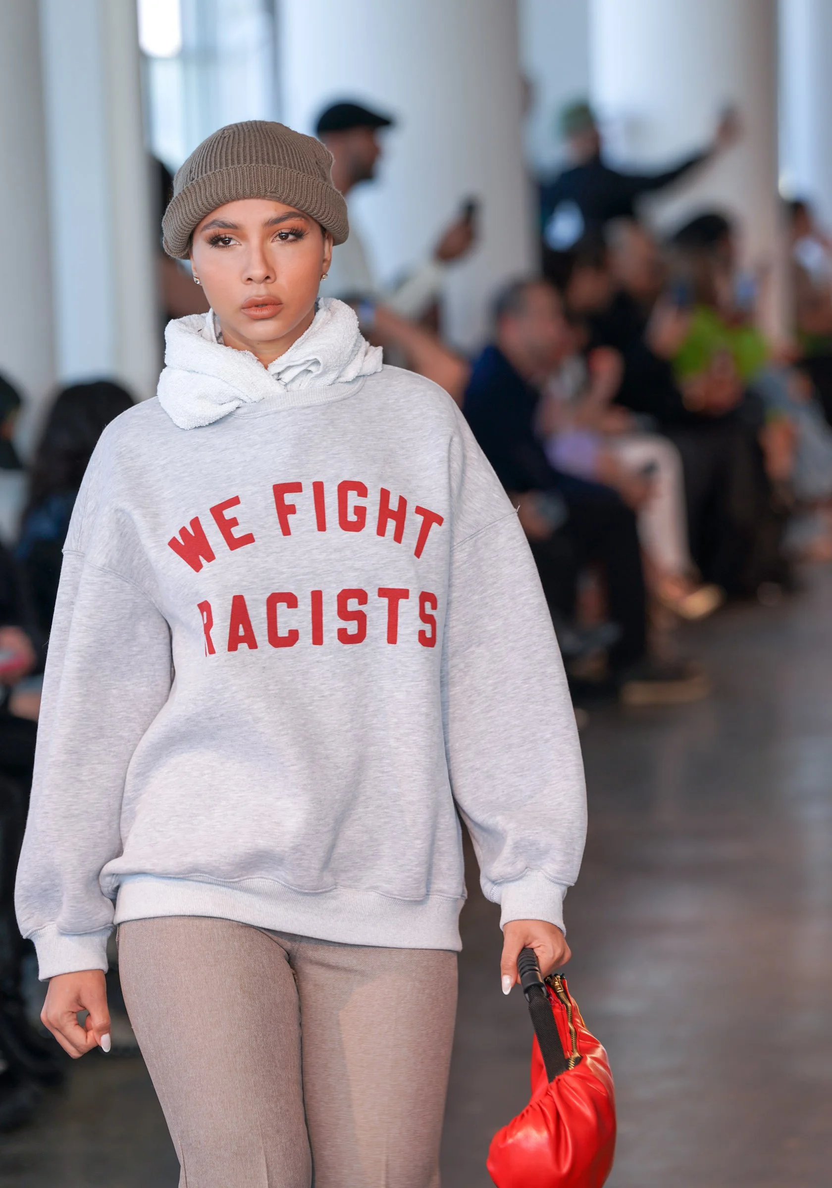 A woman in casual clothing walking in an indoor space, carrying a red bag in her right hand. She is wearing a gray hoodie with red text that says 'We Fight Racists,' a beige beanie, and beige pants. There are people in the background sitting and stan