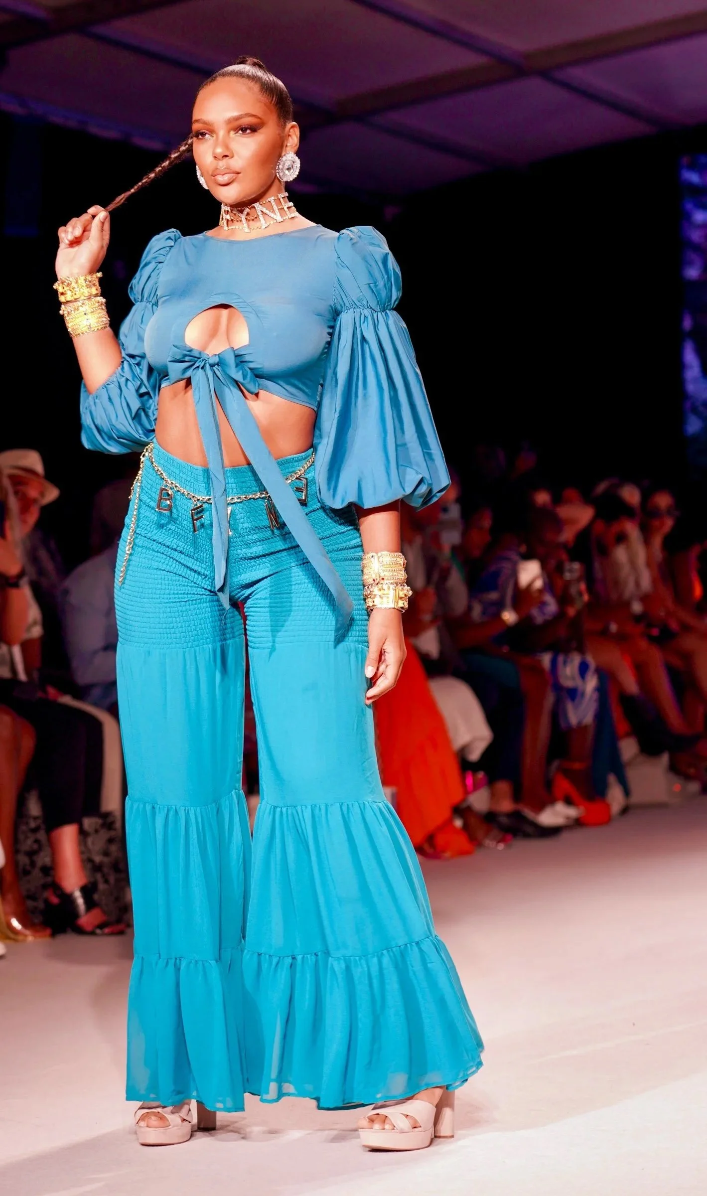 A woman walking on a runway, wearing a bright blue two-piece outfit with ruffled wide-leg pants and a top with puffed sleeves and a tied front, accessorized with large earrings, a choker, and gold jewelry, holding a strand of her hair.
