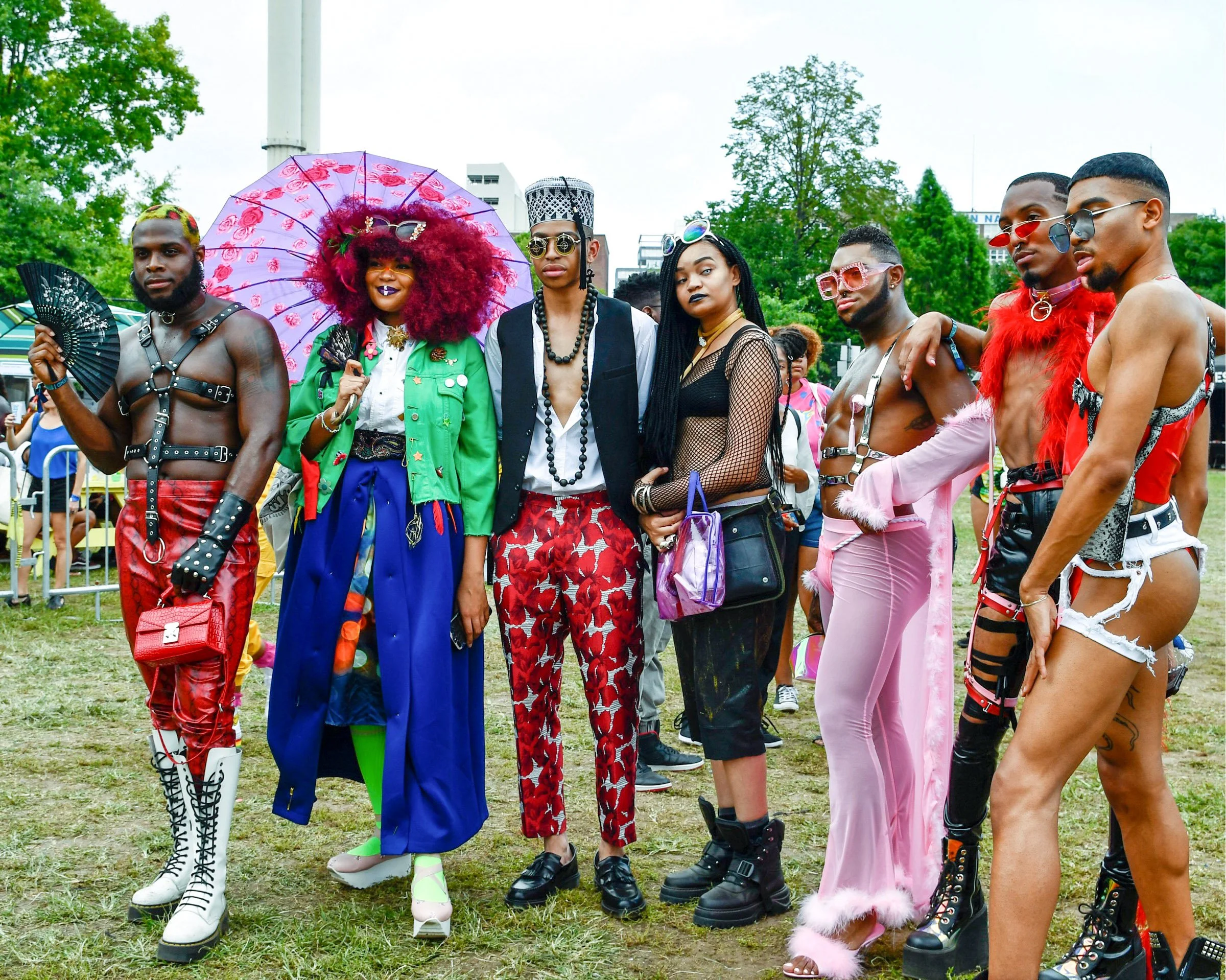 Group of diverse people dressed in colorful and flamboyant outfits, some with accessories like sunglasses and fans, standing outdoors in a park or event setting.