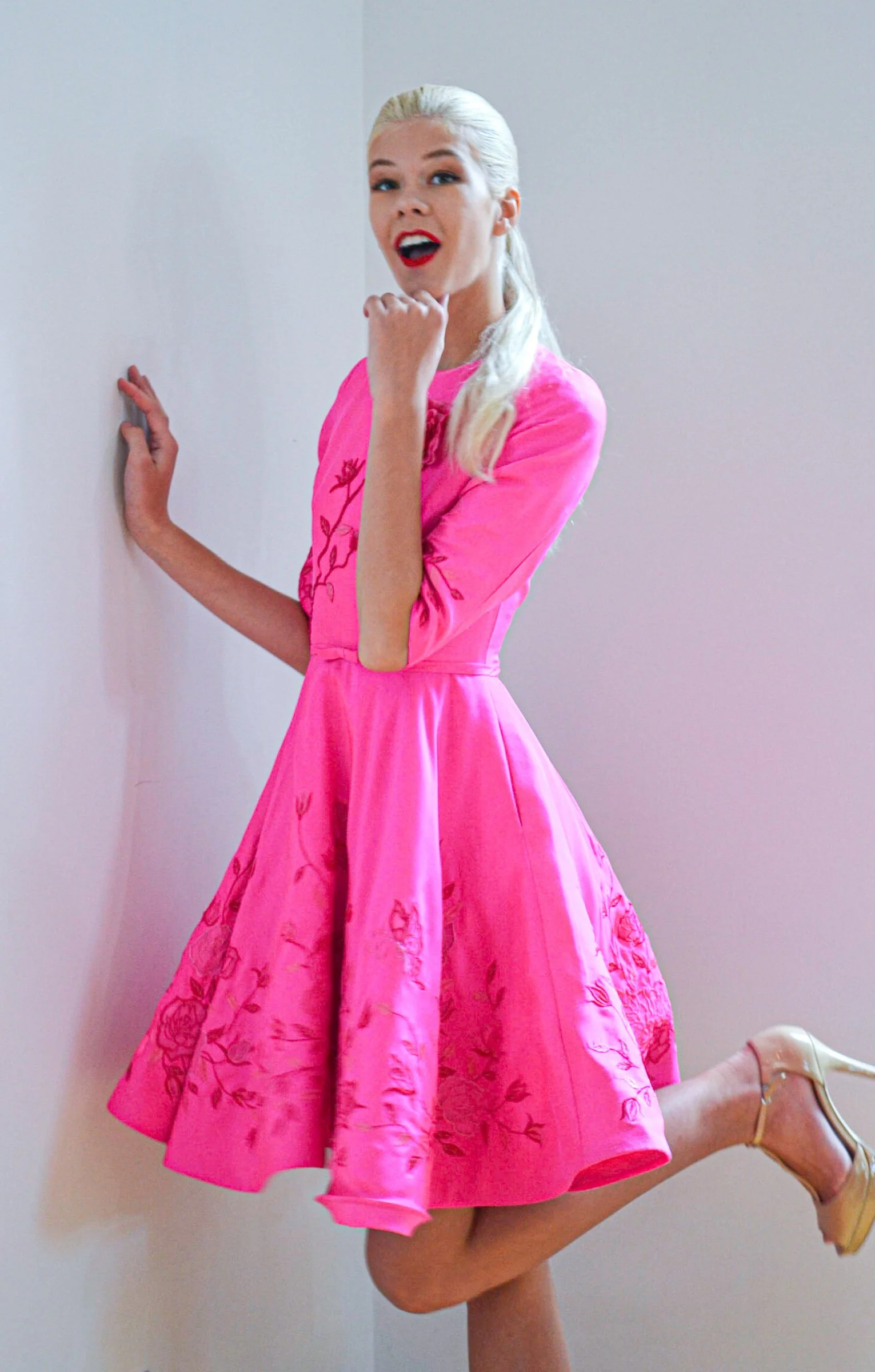 A woman in a pink dress with floral embroidery poses against a plain wall, with one leg lifted and wearing beige high heel shoes.