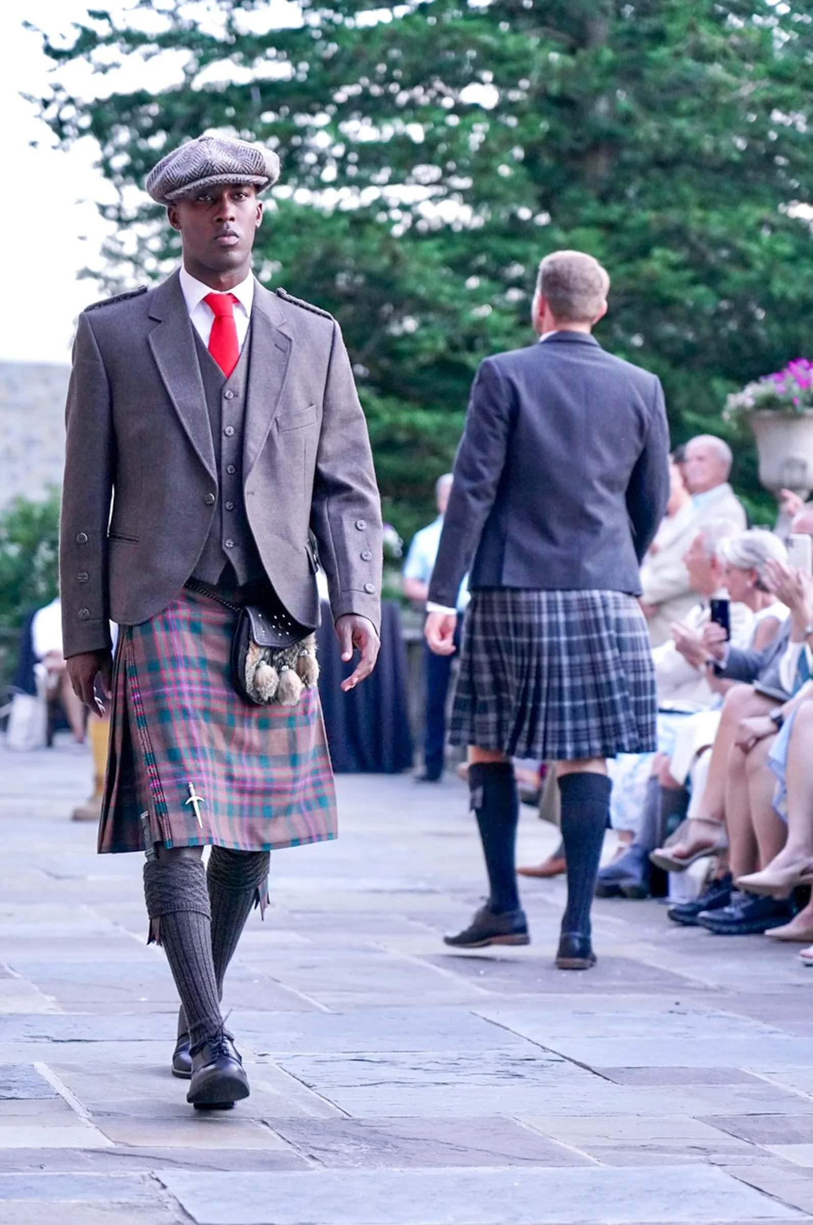 A man in traditional Scottish attire walking outdoors with a crowd watching seated on side. Background features trees and greenery.