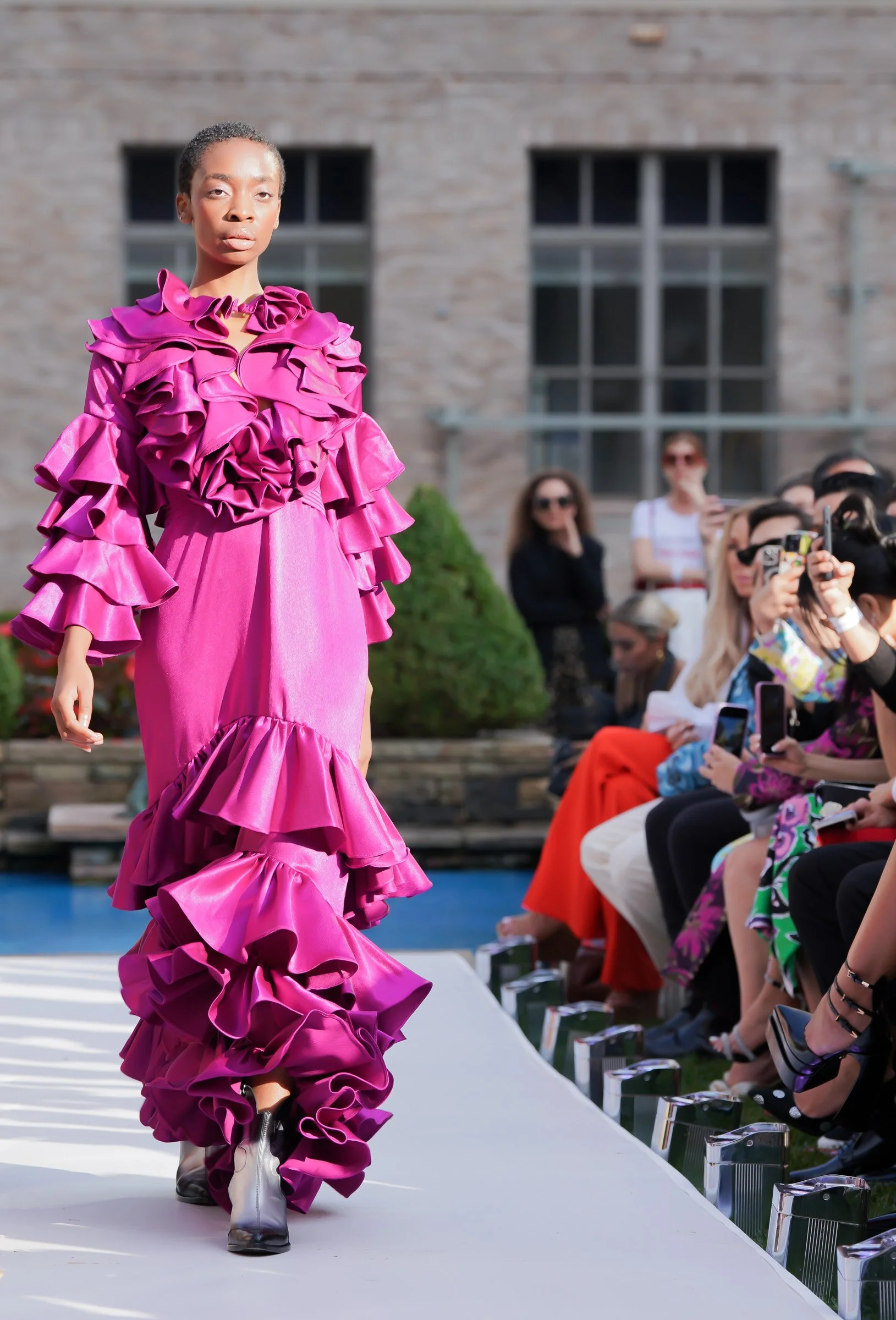 A model for Carlos Pineda walking down an outdoor runway during a fashion show, wearing a vibrant pink dress with multiple ruffles and ruffled sleeves, and white ankle boots. Audience members sit along the runway, some taking photos.
