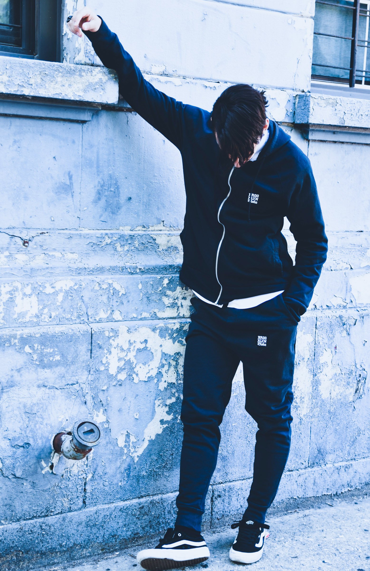 A person leaning against a weathered concrete wall, wearing a black hoodie, black pants, and black and white sneakers. Their head is bowed, and one arm is raised above their head, touching the wall.