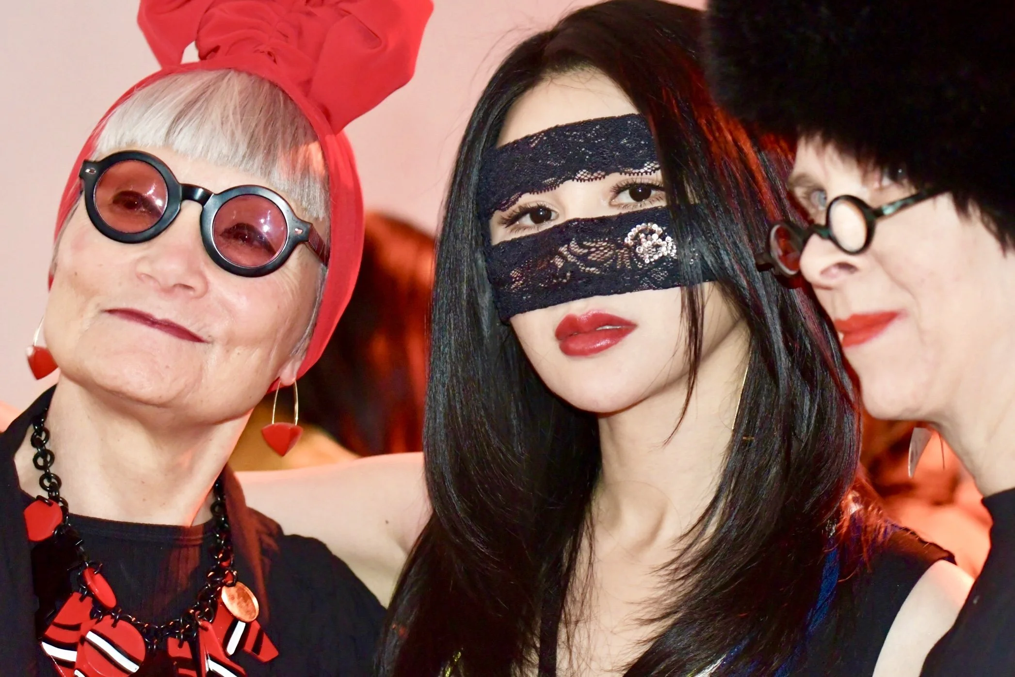 Three women dressed in alternative fashion, with the woman on the left wearing sunglasses, a red headwrap, and bold makeup, the woman in the middle wearing a lace mask covering her eyes and nose, with dark hair and makeup, and the woman on the right 