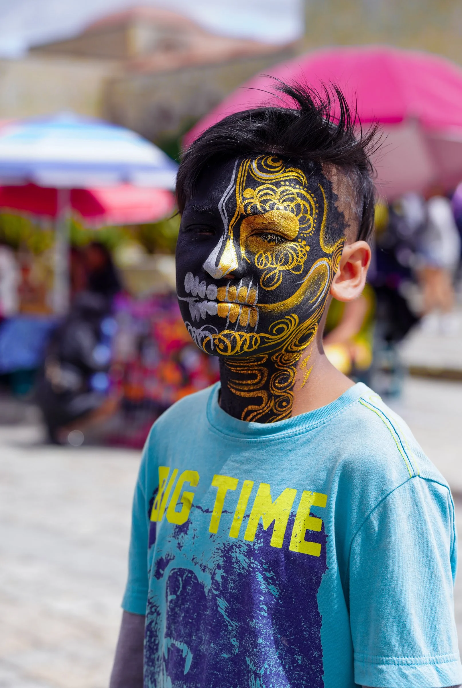 A boy with his face painted as a black and yellow skull, wearing a light blue t-shirt with a graphic design and the words 'Big Time.' He has pink hair and is outdoors with colorful umbrellas in the background.