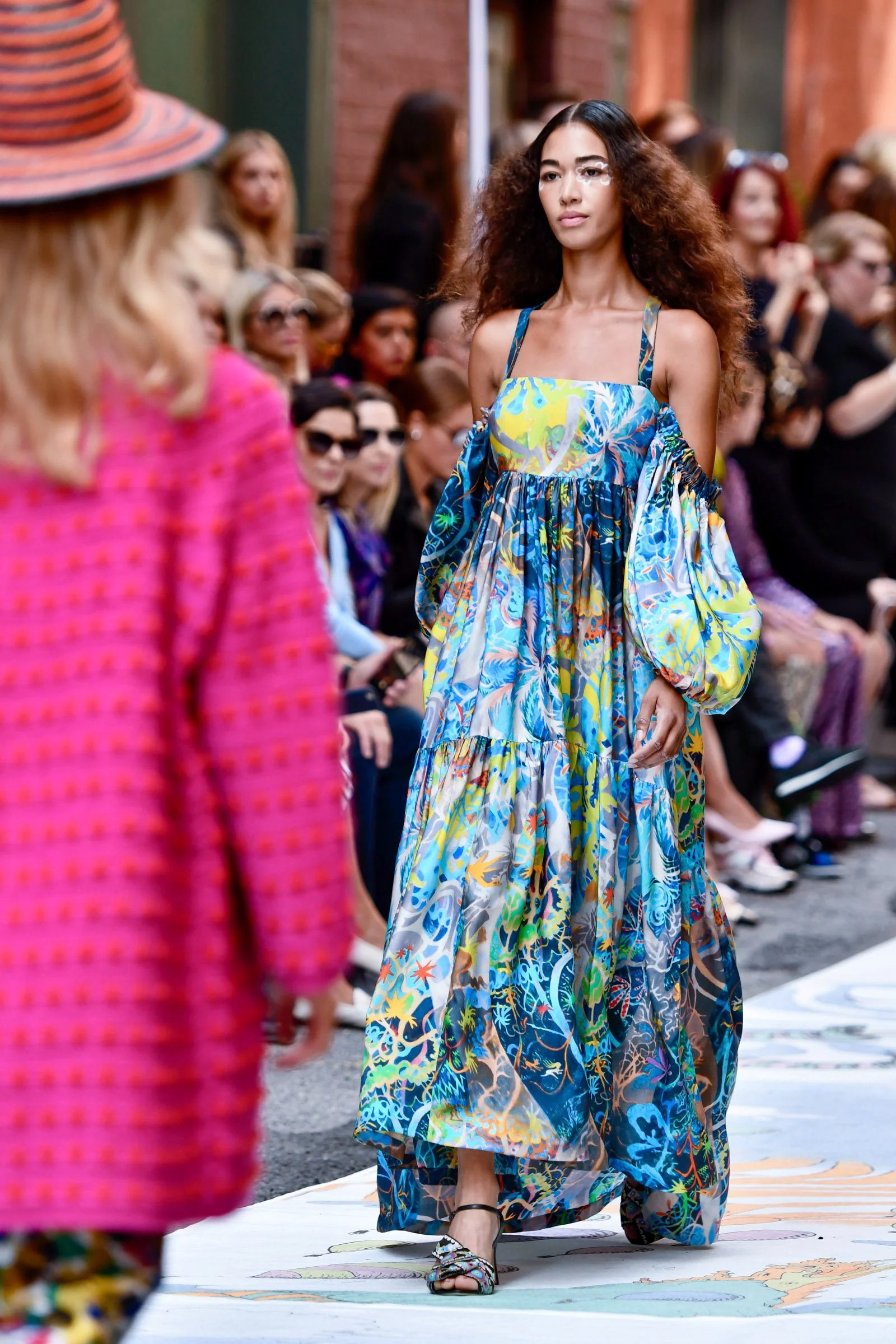 A woman with curly hair and floral eyeliner walking down a runway at an outdoor fashion show, wearing a colorful, patterned dress with a flowing skirt. Designer, Cynthia Rowley.