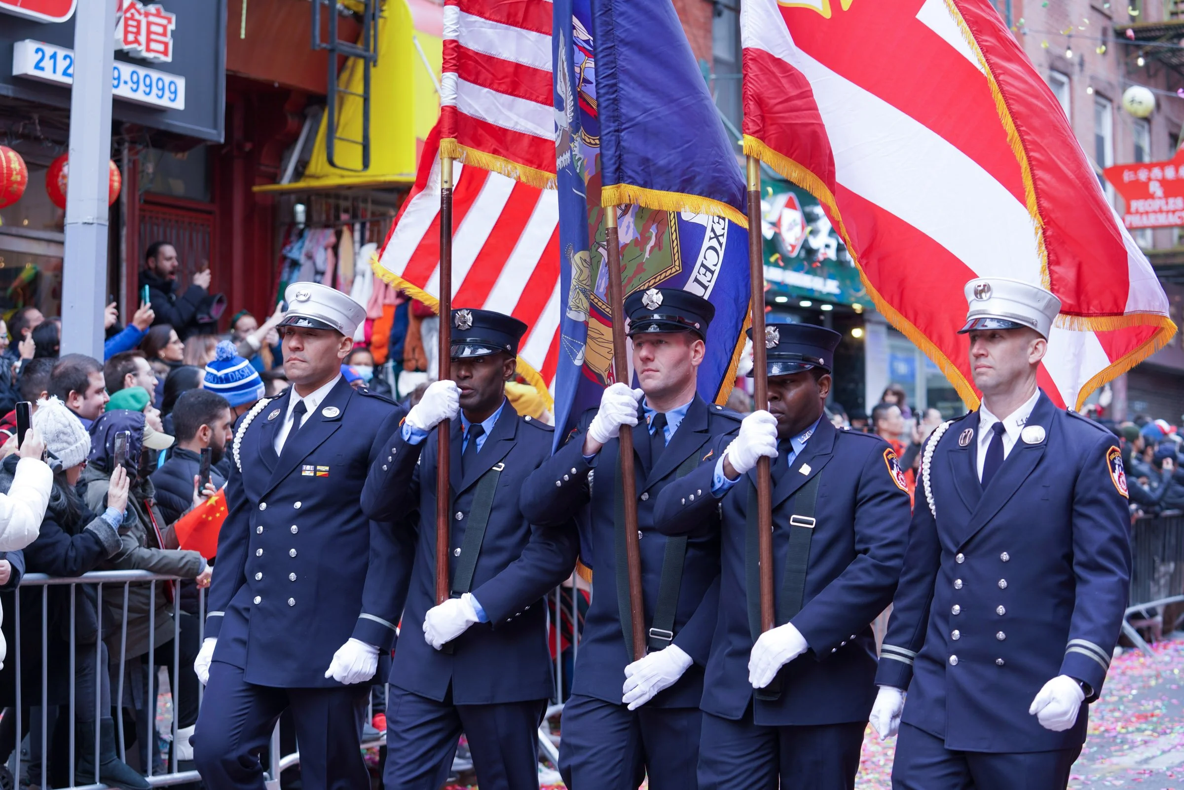 Four uniformed individuals holding flags participate in a parade, with spectators behind barricades on a city street decorated for celebration. NY Chinese New Year Parade.