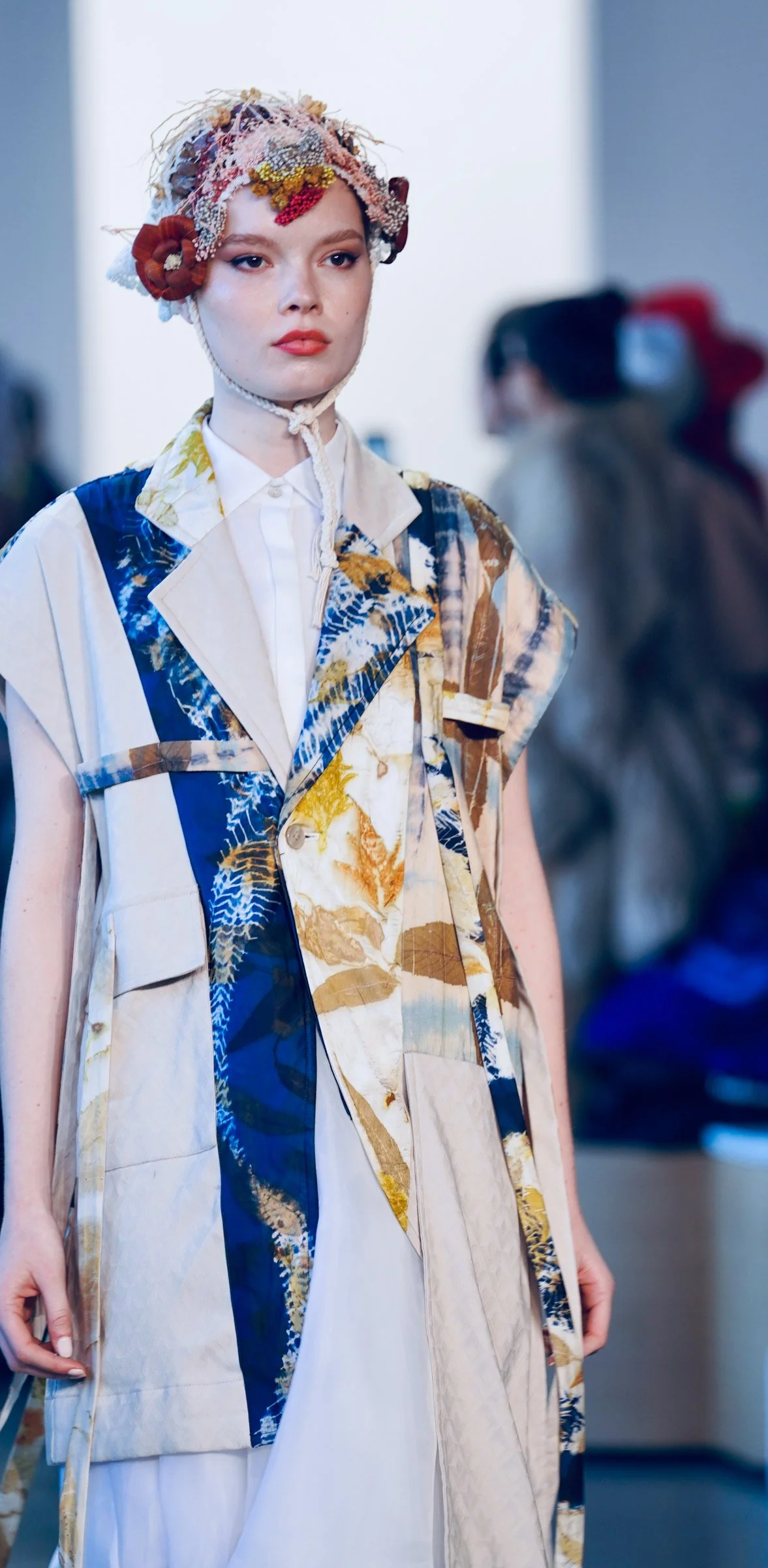 A woman wearing a patchwork vest with various patterns and colors, white shirt, and an intricate textured headpiece with flowers and beads.
