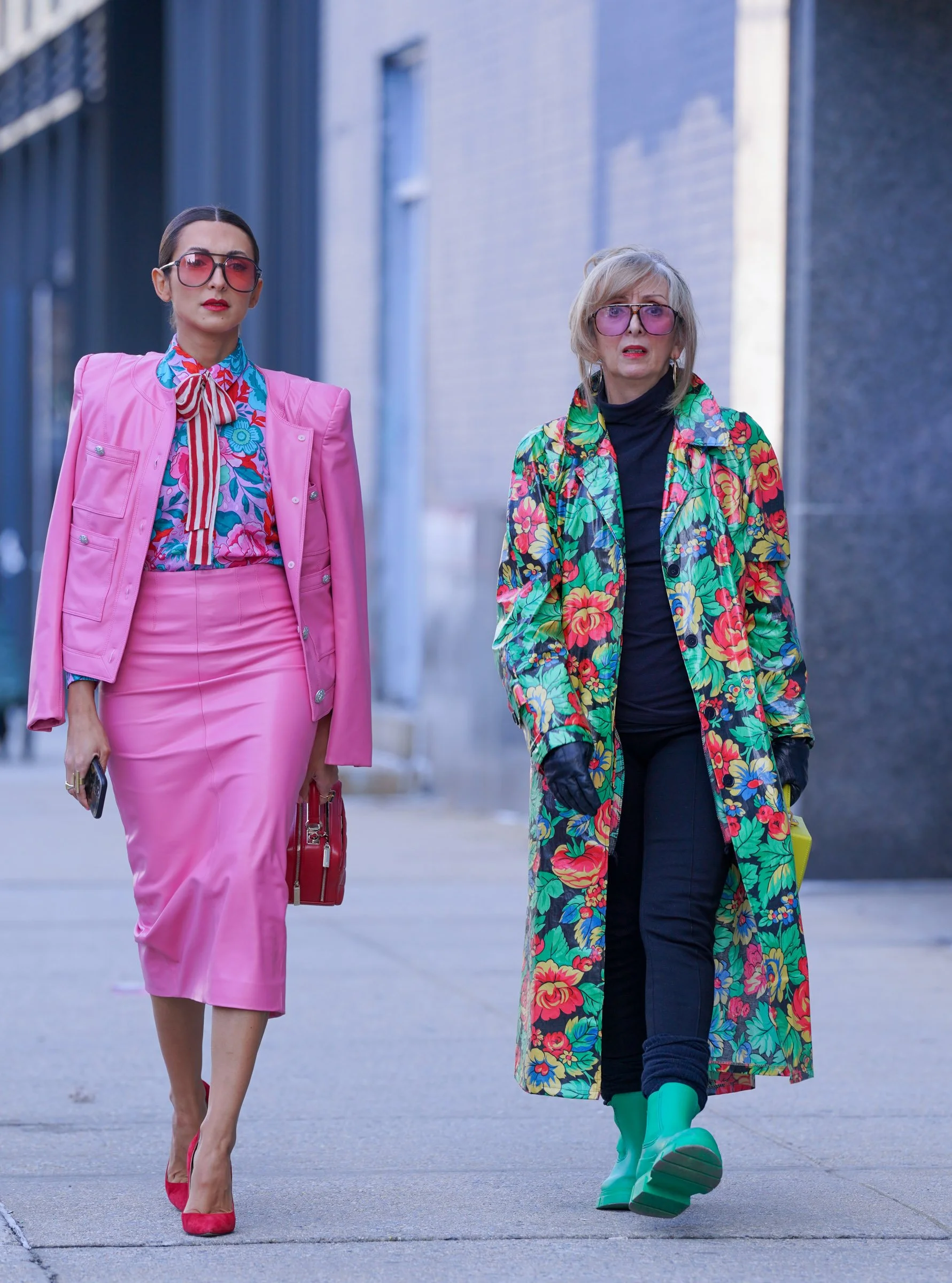 Two women walking down a city street, one in a bright pink outfit with sunglasses, another in a colorful floral coat with turquoise boots.