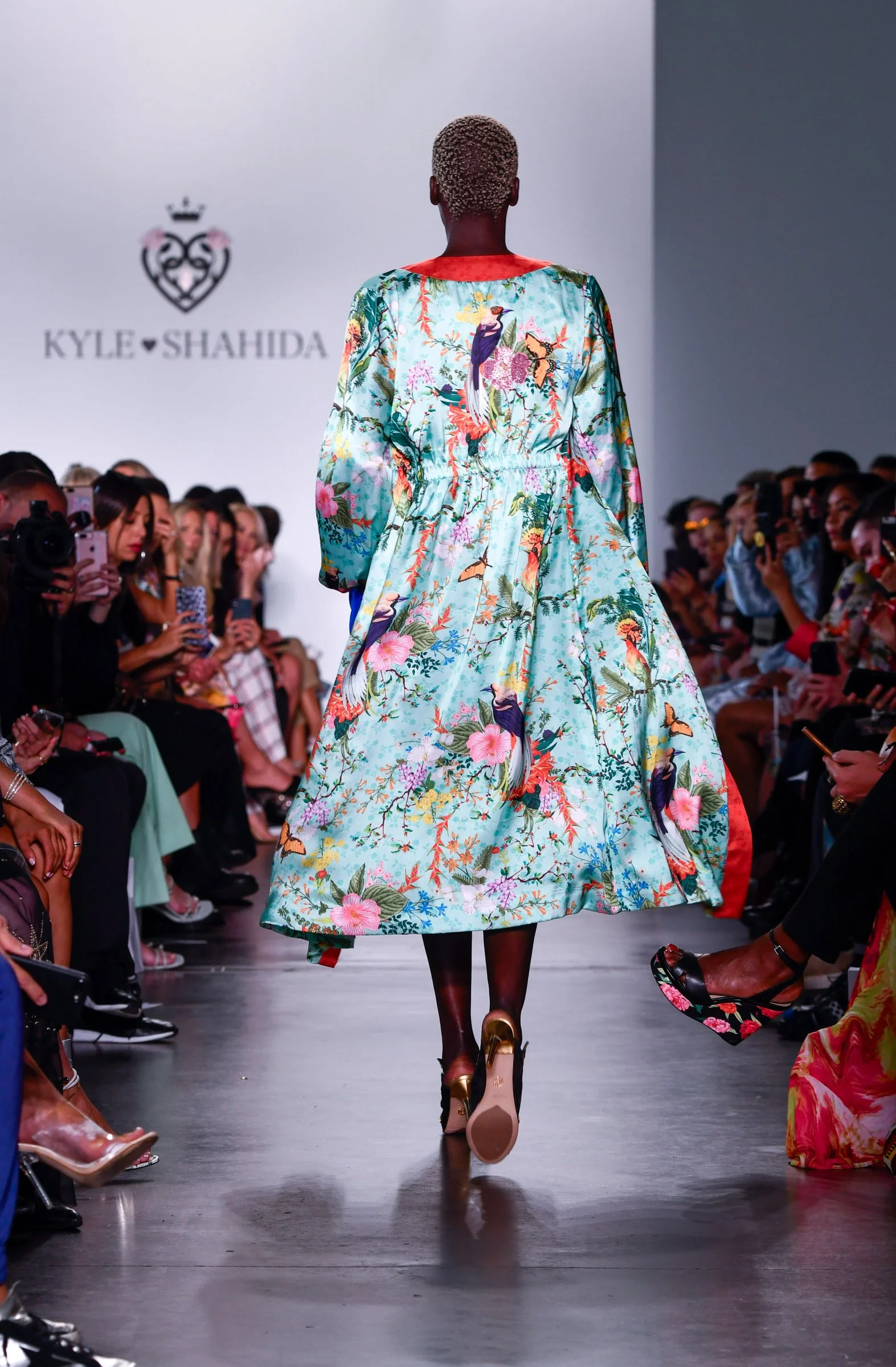 Model walking down runway during a fashion show featuring a vibrant floral and bird patterned dress.