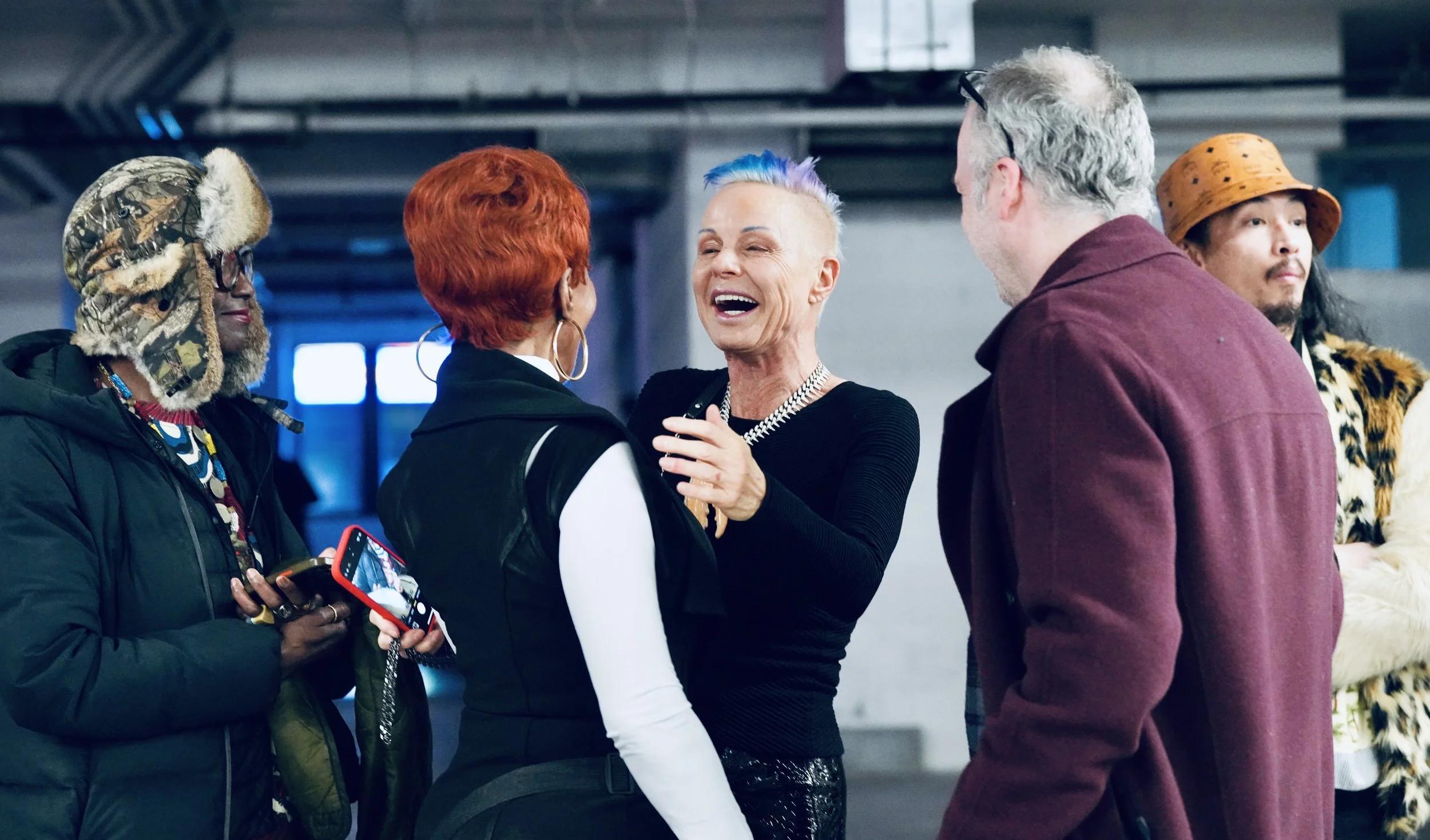 A group of diverse people socializing indoors, with one woman with multicolored hair laughing in the center, surrounded by others in stylish outfits.