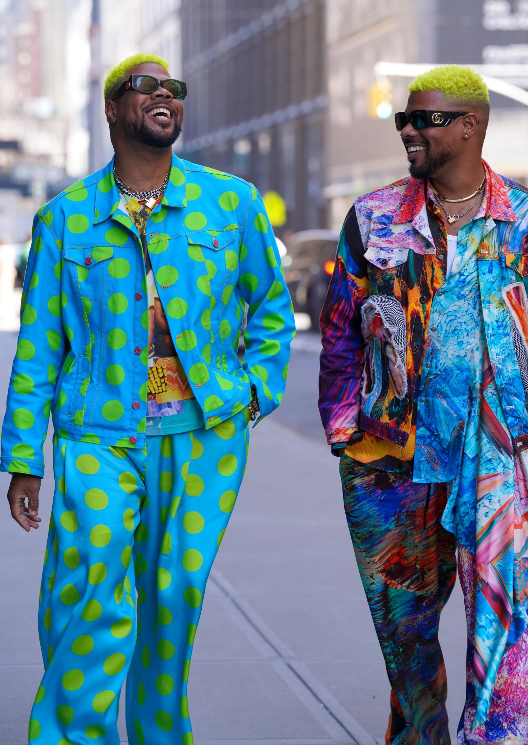 Two men walking on a city street, smiling and wearing colorful, patterned clothing and sunglasses. One man has bright yellow hair and the other has green hair.