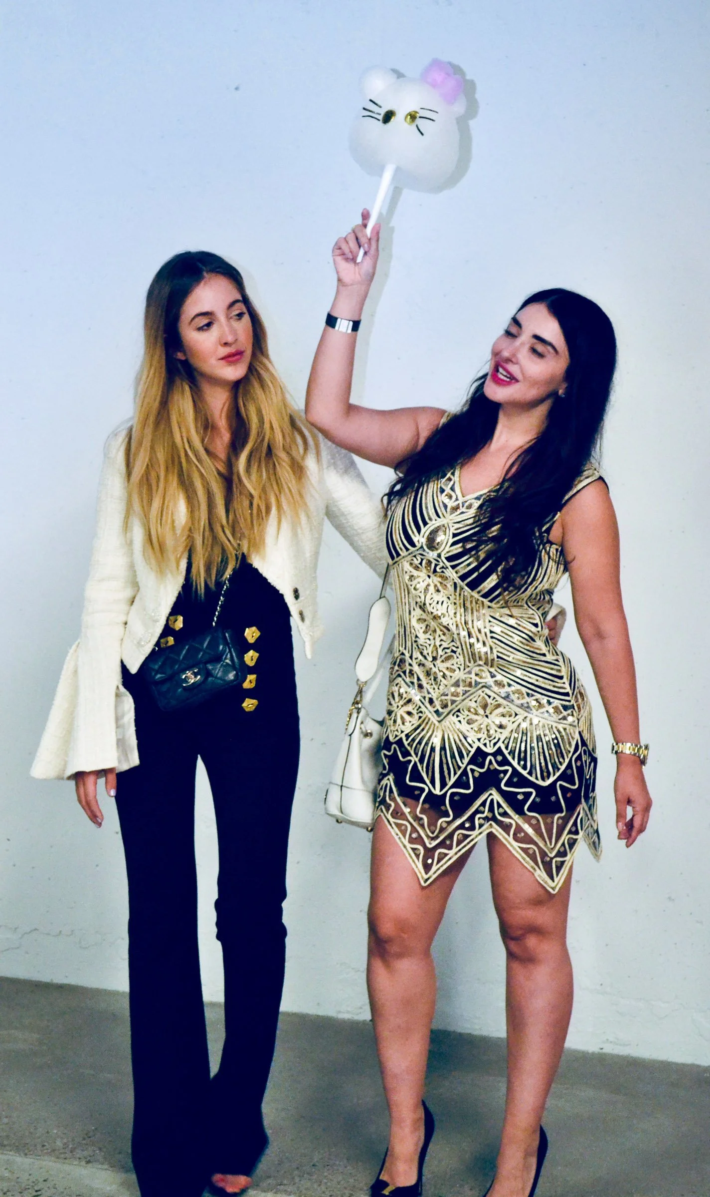 Two women standing against a plain wall, one holding a Hello Kitty-themed cotton candy on a stick. The woman on the left has long blonde hair, wearing a white blazer over black clothing, and has a black purse with gold accents. The woman on the right