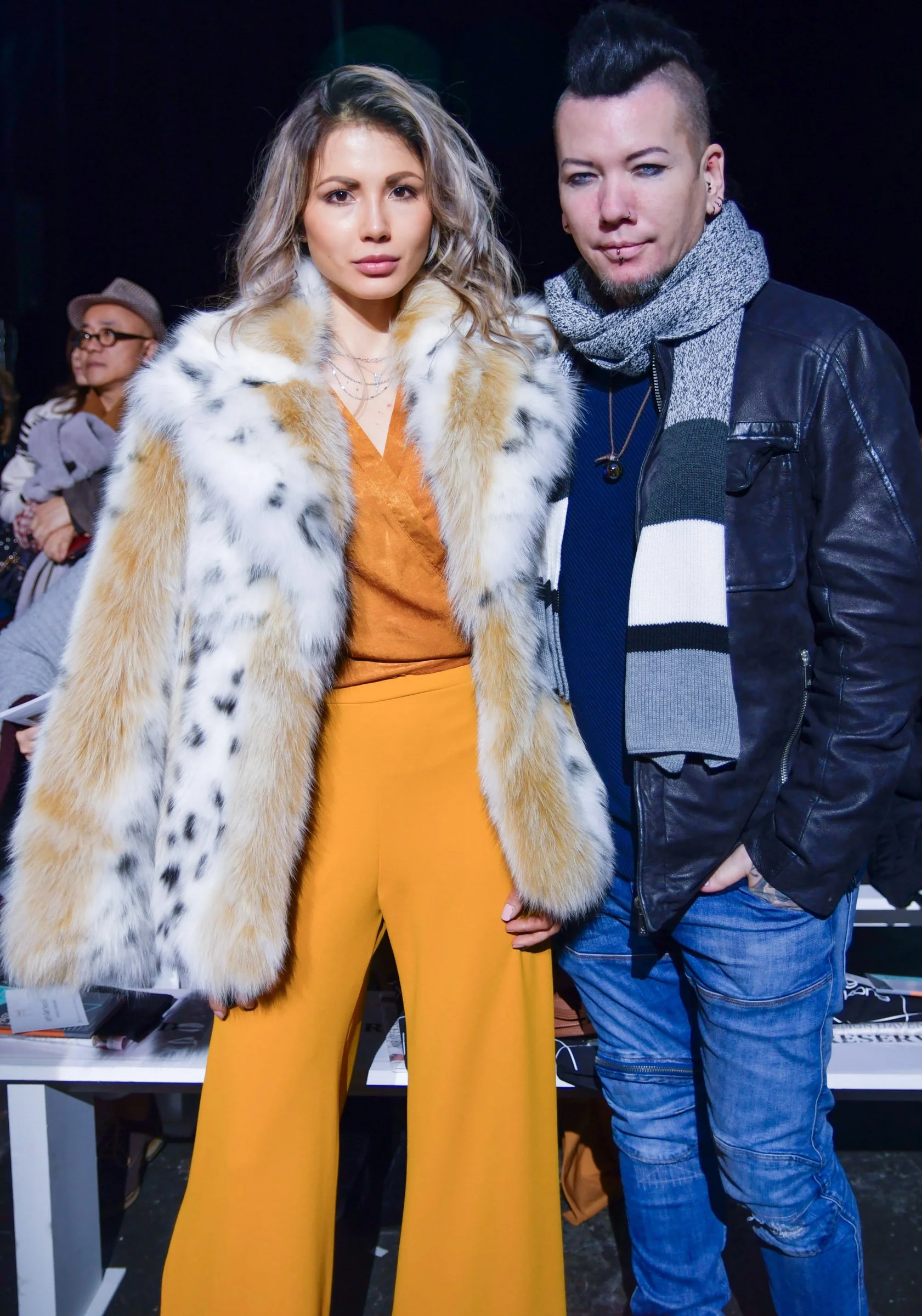 Two people standing together, one woman wearing a fur coat and yellow pants, the other man in a leather jacket and scarf, at an indoor event with people in the background.