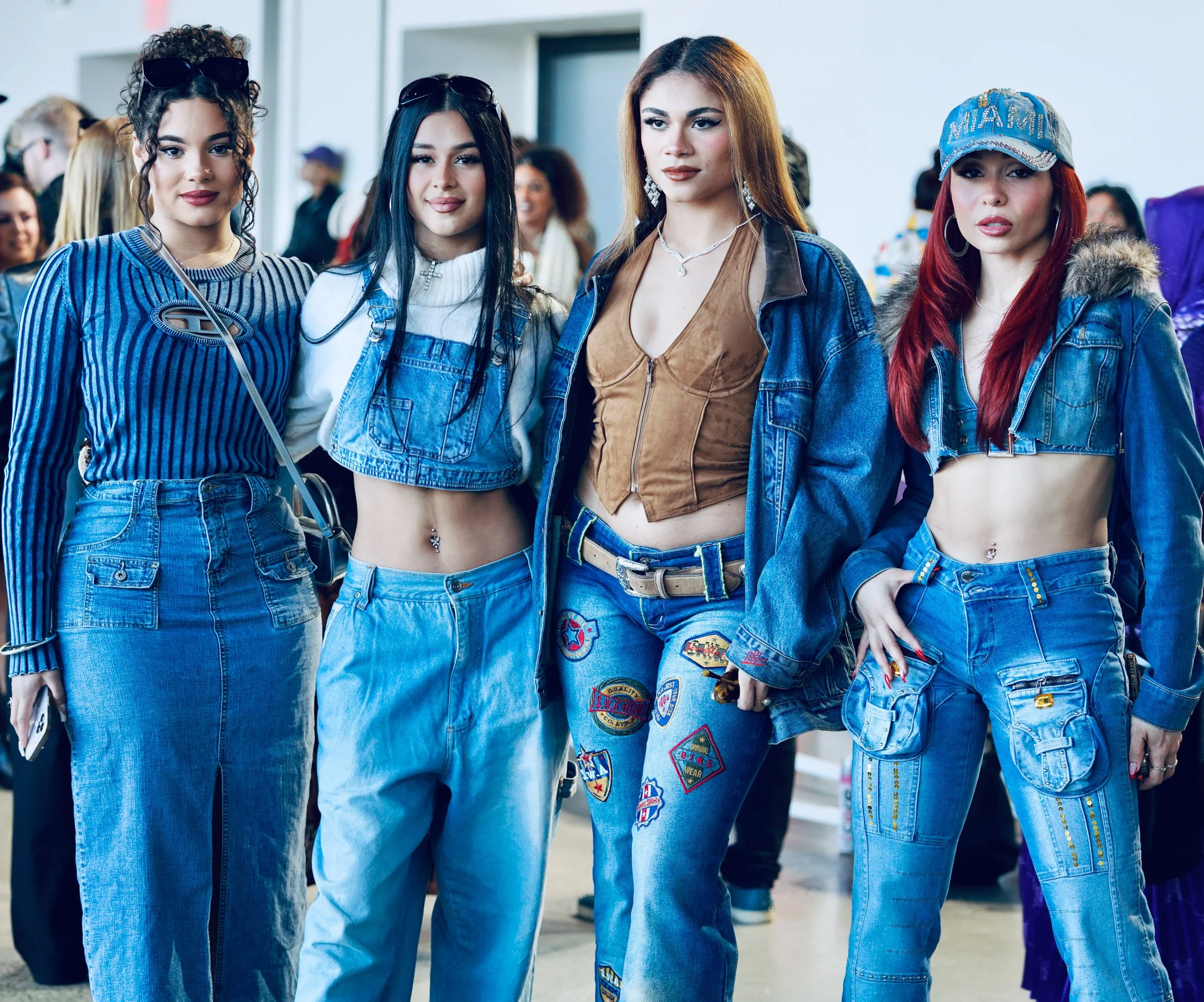 Group of four diverse women standing together in an indoor setting, dressed in casual denim outfits, with some wearing accessories like sunglasses, jewelry, and a Miami cap, looking confident and stylish.