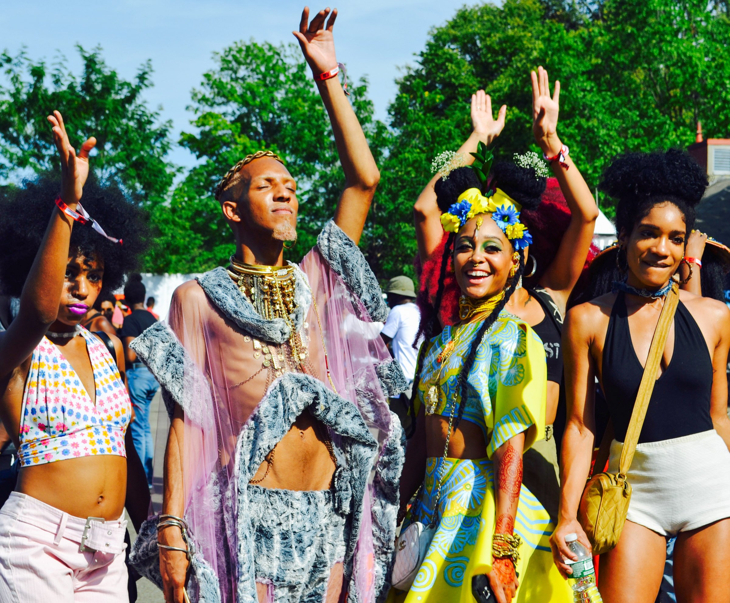 A diverse group of people participating in an outdoor celebration or festival, with some raising their hands and smiling, dressed in colorful clothing and accessories, with trees and a clear sky in the background.