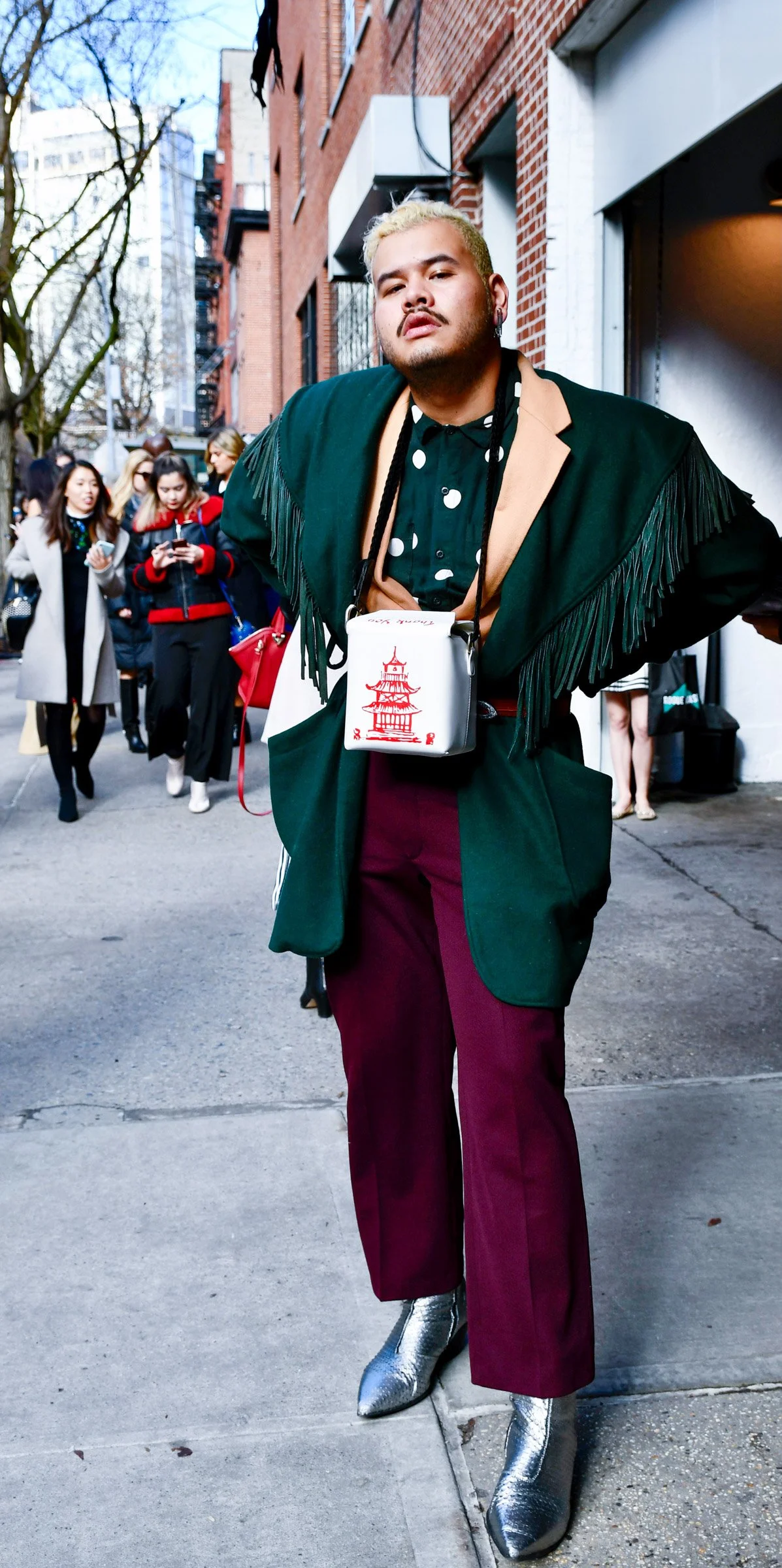 A confident person with bleached blonde hair and earrings poses on a city street, wearing a green jacket with fringe details, a black shirt with white polka dots, maroon pants, and shiny silver boots, with a group of people in the background.