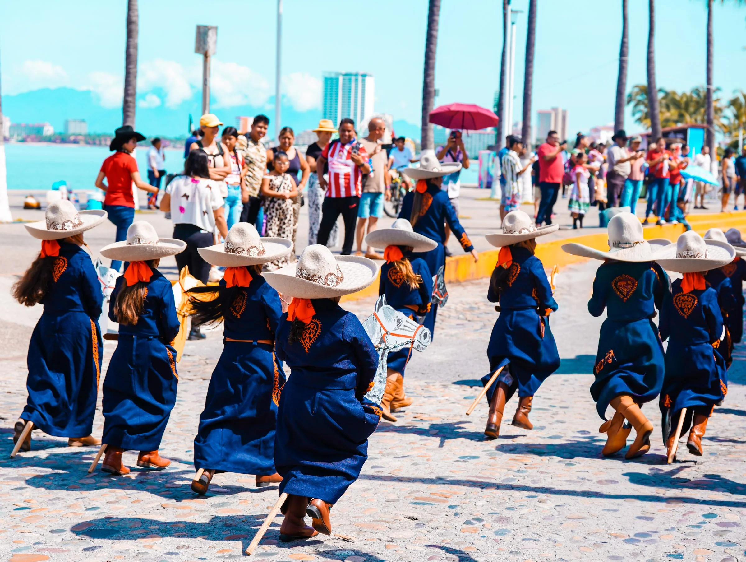People dressed in traditional Mexican clothing, including wide-brimmed sombreros, participating in a cultural parade or celebration by the waterfront. Día del Charro.
