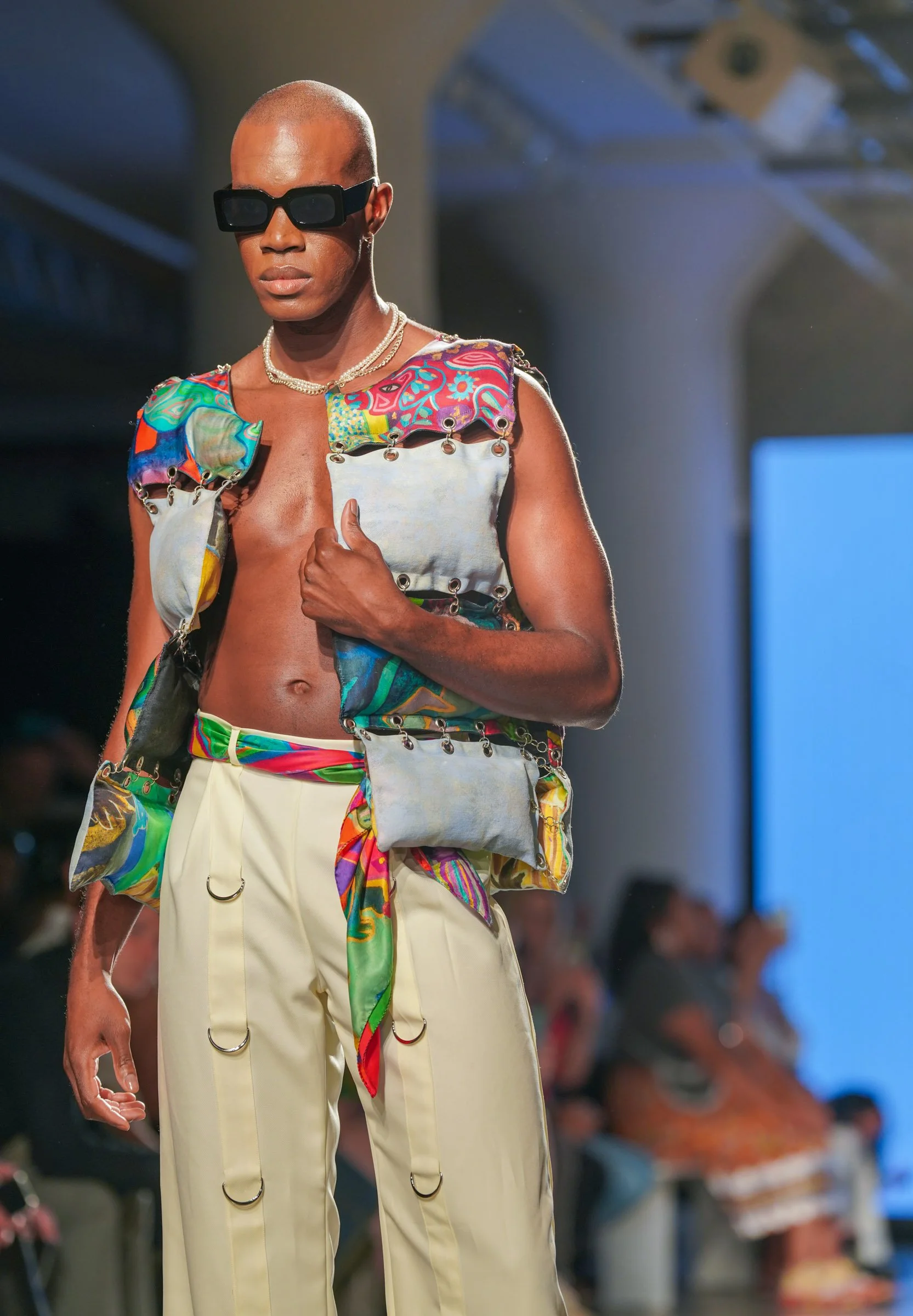 A fashion model walking on runway, wearing oversized black sunglasses, layered necklaces, and a colorful, patchwork vest with metal rings, paired with high-waisted beige pants that have metal rings and colorful fabric accents.
