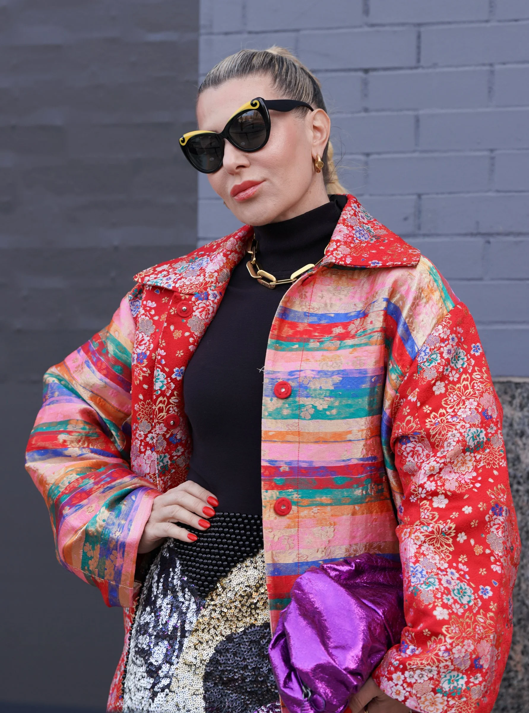 A woman wearing large black sunglasses with yellow accents, a gold chain necklace, gold earrings, a black turtleneck, a colorful patterned jacket, and holding a purple shiny handbag, stands against a gray brick wall.