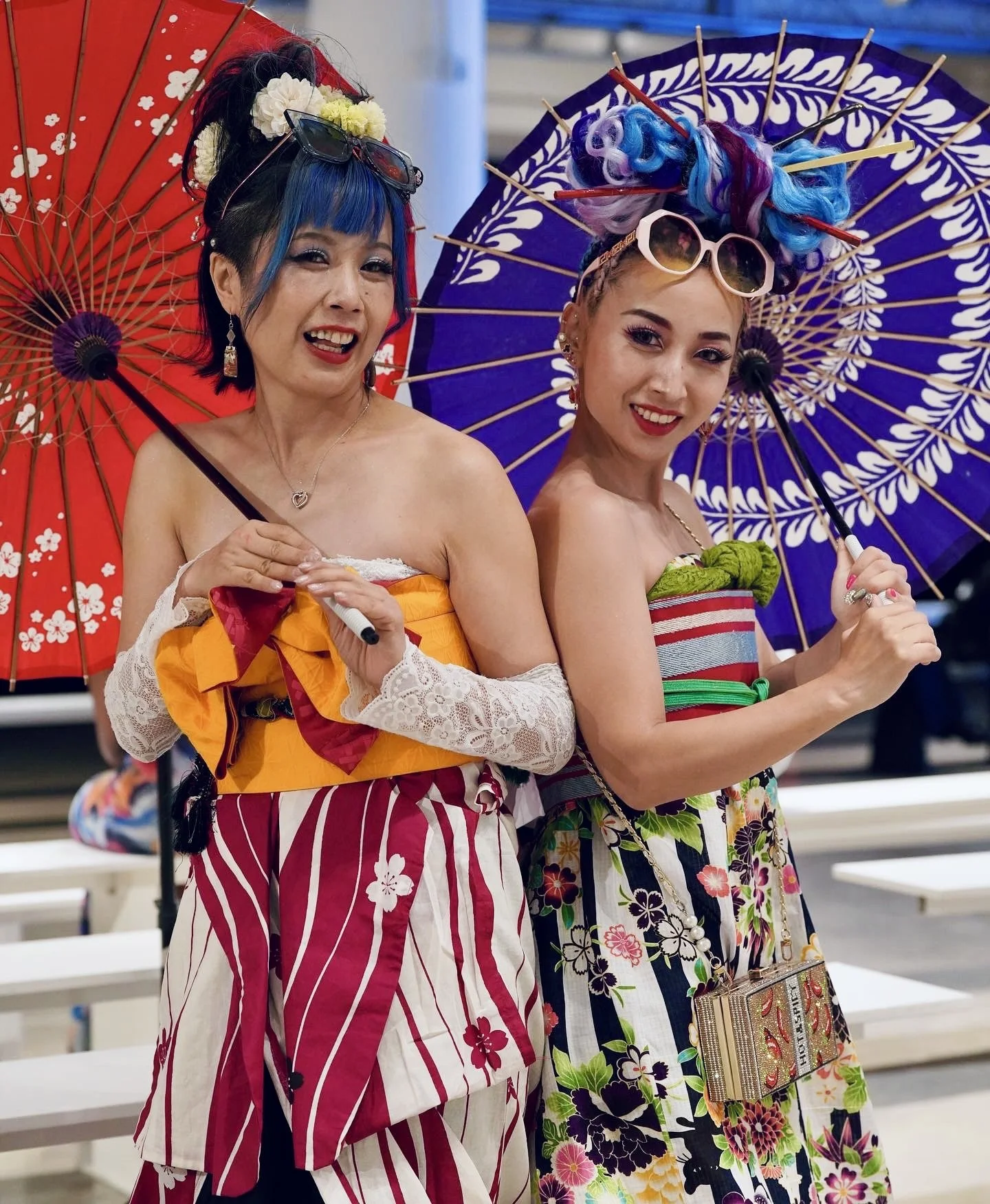 Two women holding parasols at a daytime outdoor event, wearing colorful summer dresses with floral patterns. The woman on the left has short blue hair with glasses on her head, while the woman on the right has brightly colored curly hair with sunglas