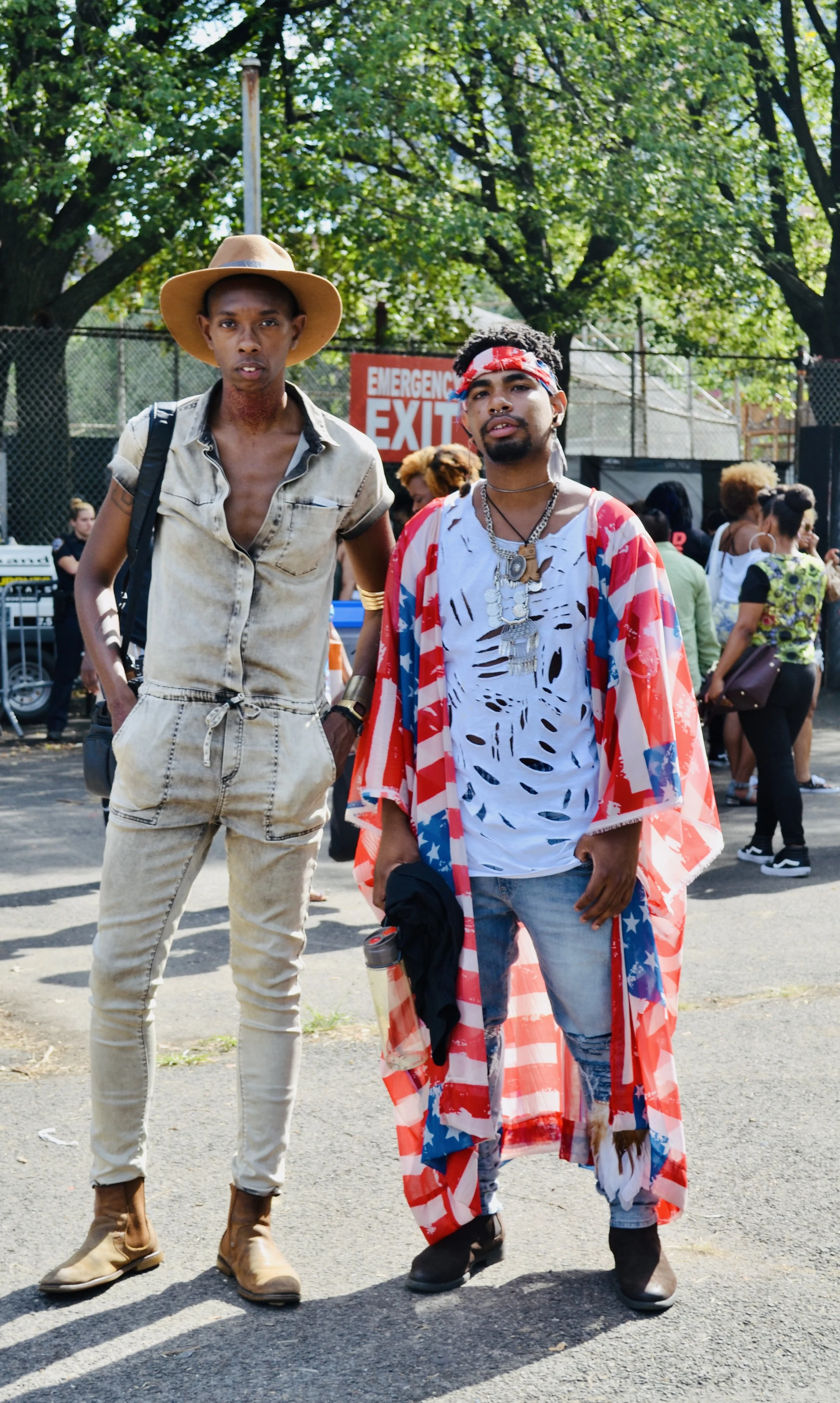 Two young men standing outdoors in a park with trees and a crowd in the background. One wears a wide-brimmed hat and beige jumpsuit, the other wears a colorful cape, a red bandana, and layered necklaces.
