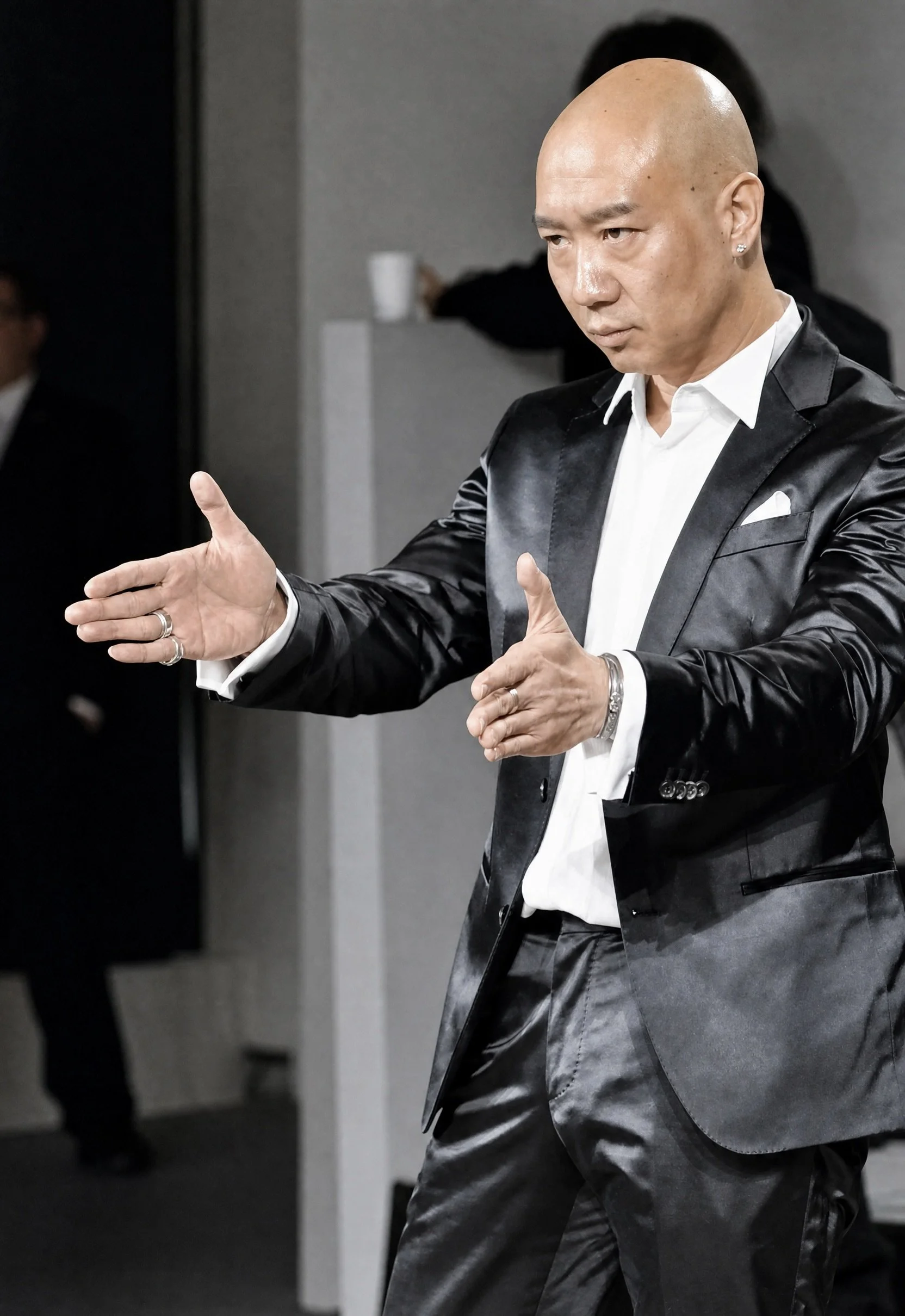 A man in a shiny black suit with a white shirt is standing indoors with his hands extended outward. He has a shaved head and an earring in his left ear. There is a person in the background holding a cup.