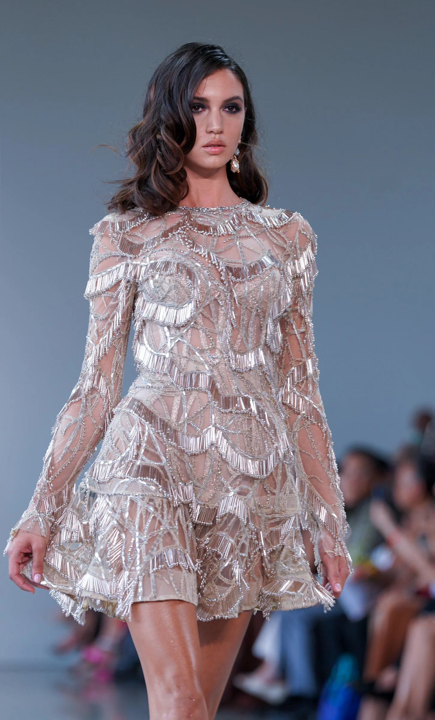 A female model walking on a runway wearing a sheer, beaded dress with intricate silver embellishments and earrings.Indonesia Now show during NYFW 2024.