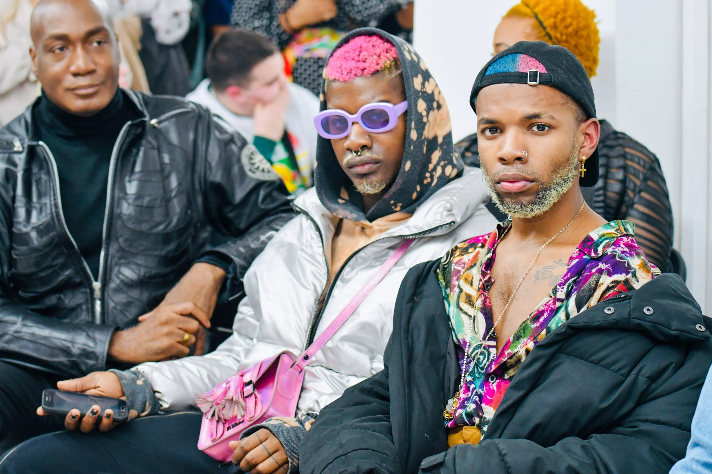 Three people sitting close together, with a blurred crowd in the background, wearing colorful and distinctive clothing and accessories, including sunglasses, hats, and jewelry, in what appears to be a public indoor event or gathering. 