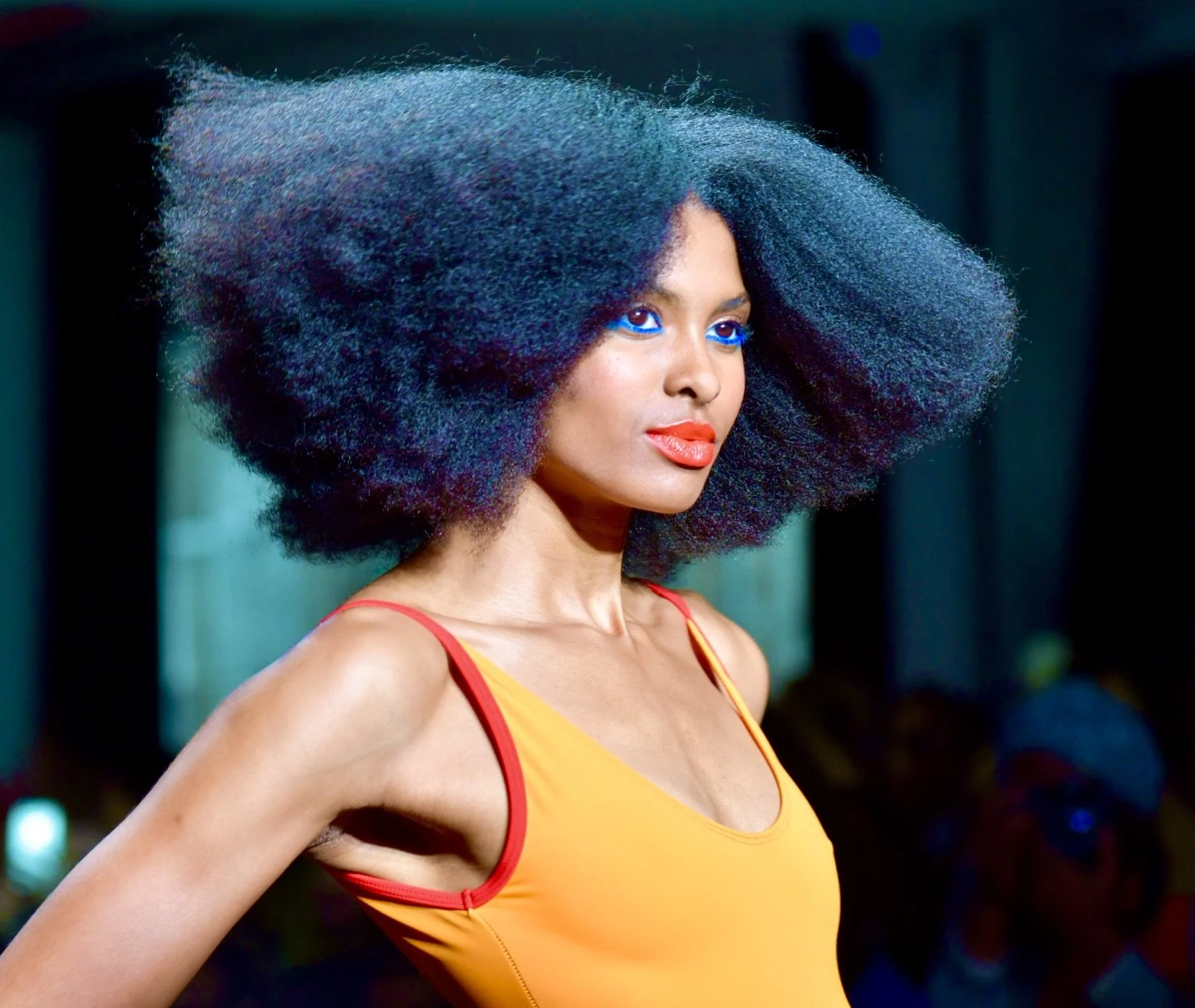 A woman with voluminous curly black hair and bright blue eyeshadow, wearing a yellow top with red straps, looking to the side. Chromat swimwear during NYFW.