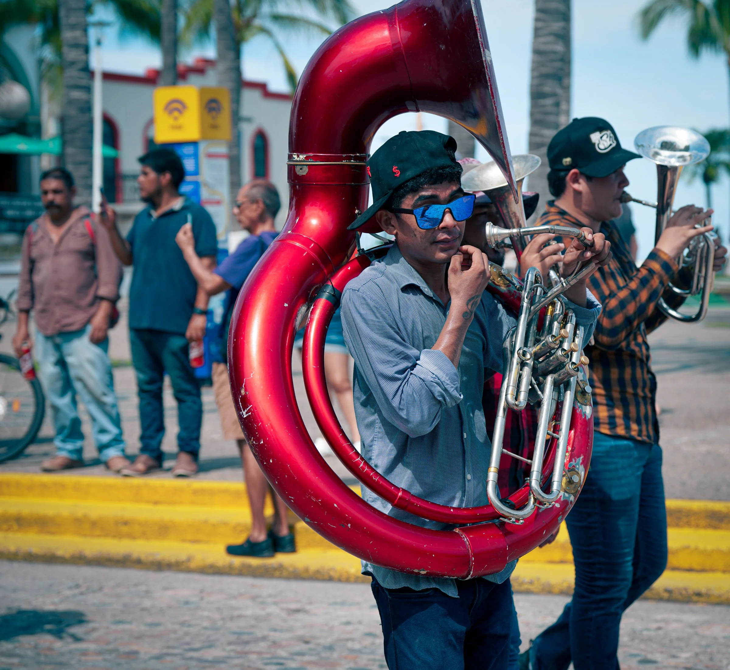 A person playing a sousaphone, part of a marching band, on a street with palm trees and onlookers in the background during daytime.
