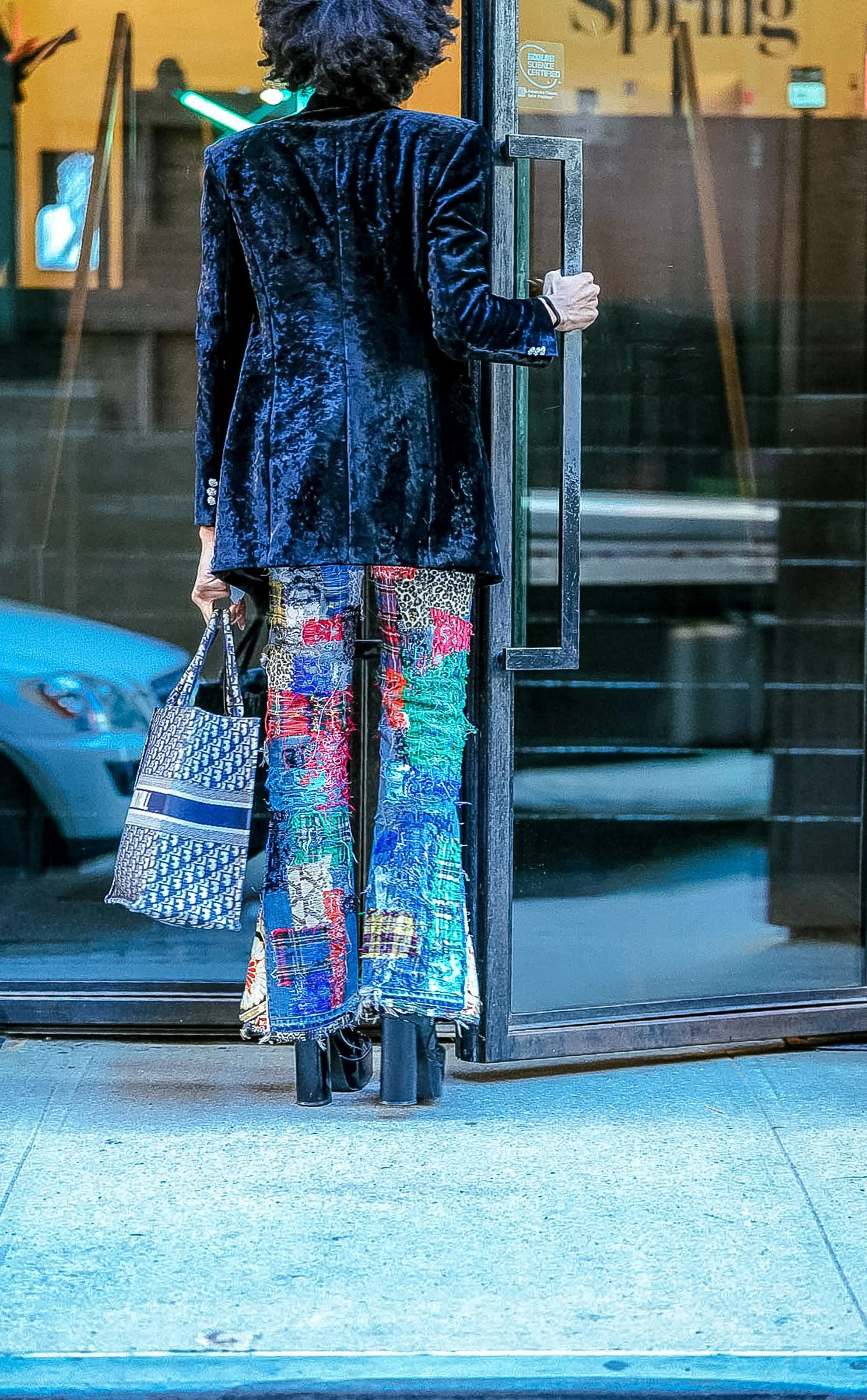 Person with curly hair wearing a blue velvet blazer and colorful patchwork jeans, holding a patterned tote bag, standing in front of a glass door at night. Spring Studios/NYFW.