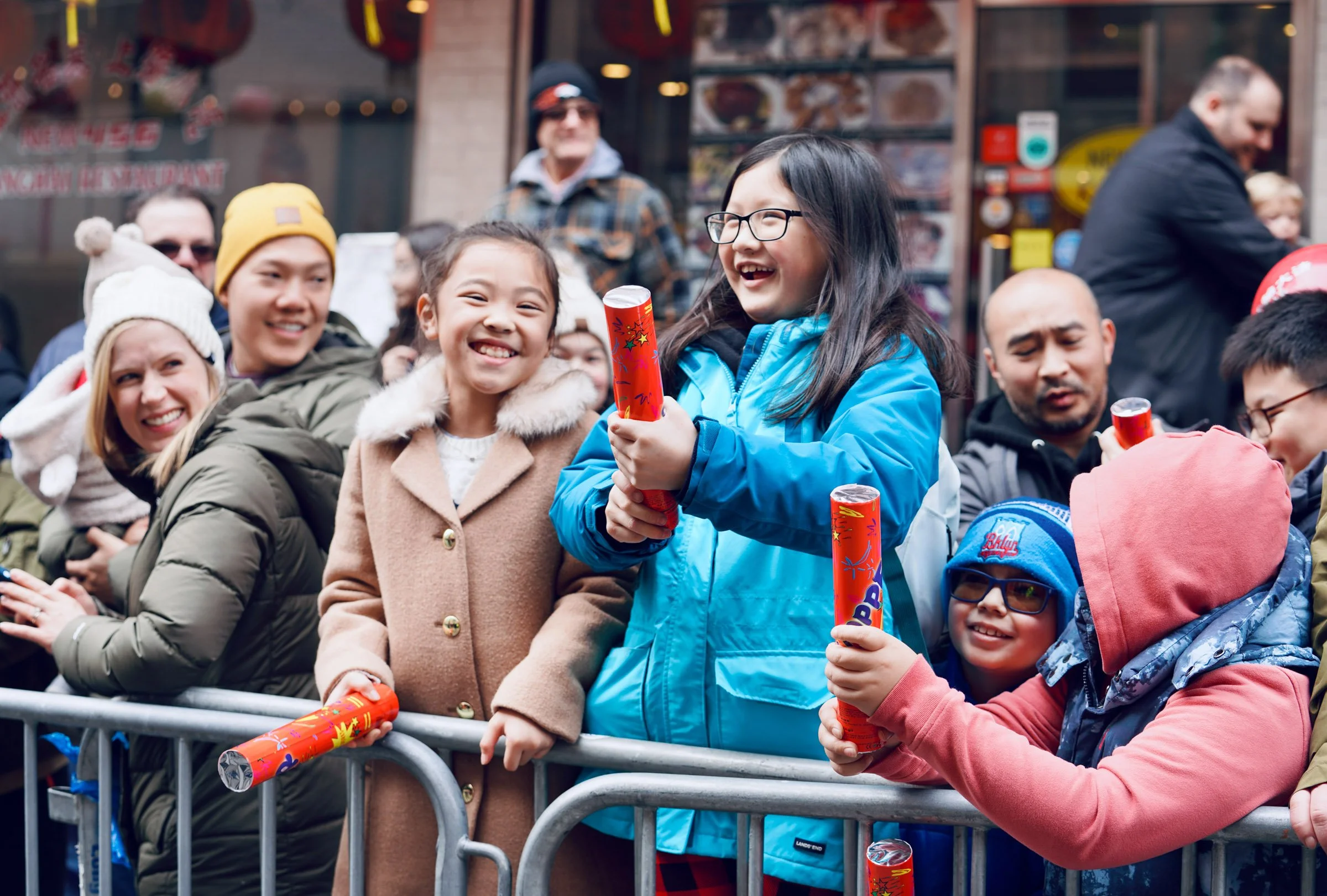Children and adults gathered behind a metal barricade, holding party poppers, smiling and celebrating outside in front of a restaurant or shop. NY Chinese New Year Parade.