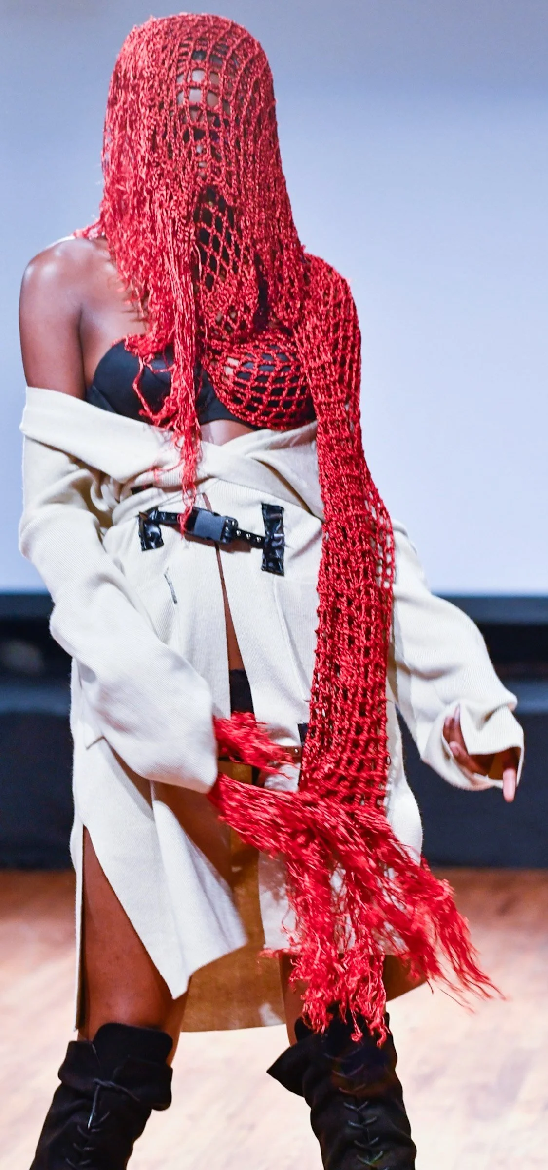 A person wearing a red, crocheted face covering, black bra, beige oversized jacket, white skirt with a high slit, and black boots.