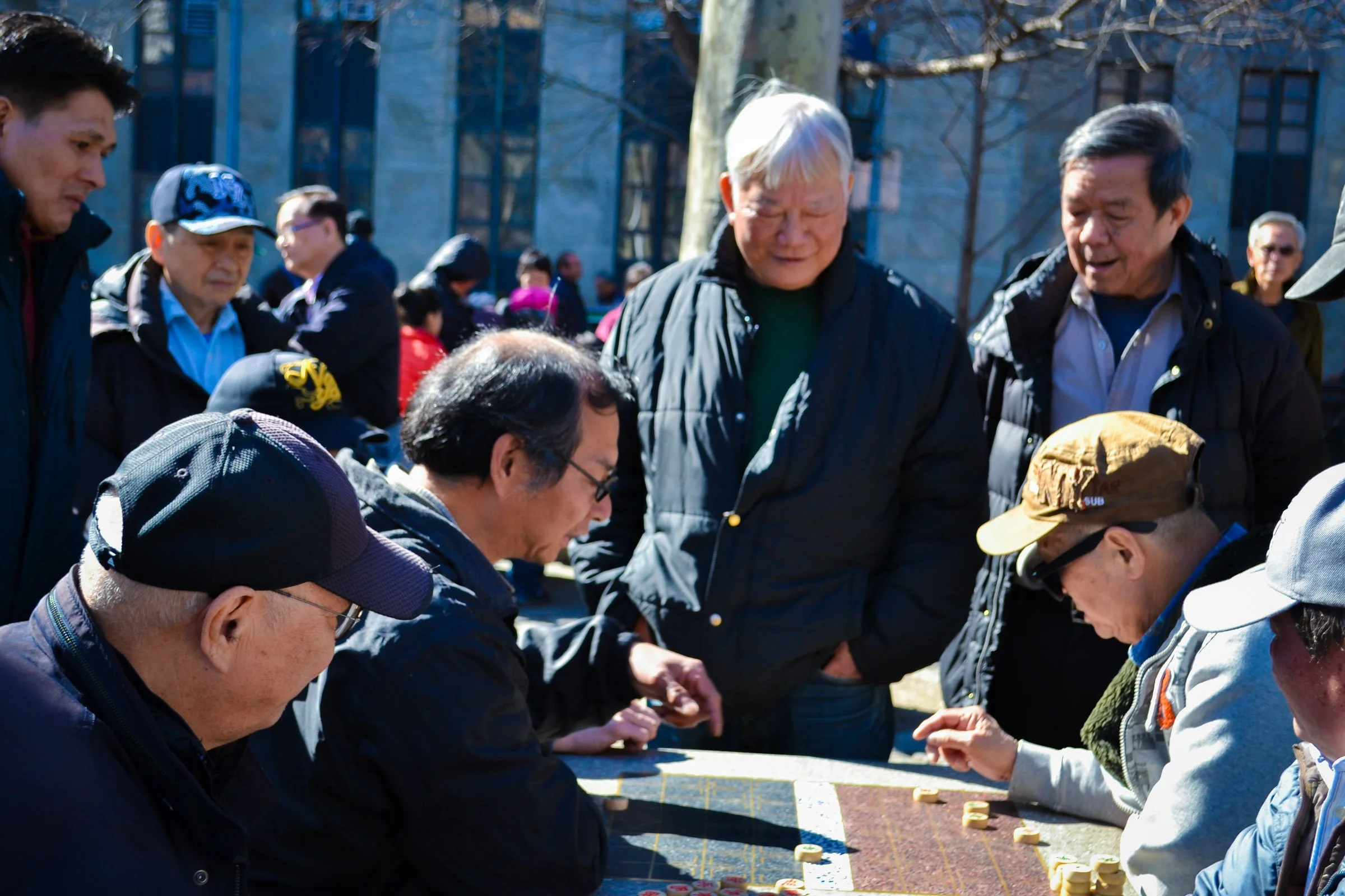 A group of people gathered outdoors on a sunny day, engaged in a game of Mahjong, with some players wearing hats and jackets, and a background of trees and a building. NYC Chinatown/Columbus Park.