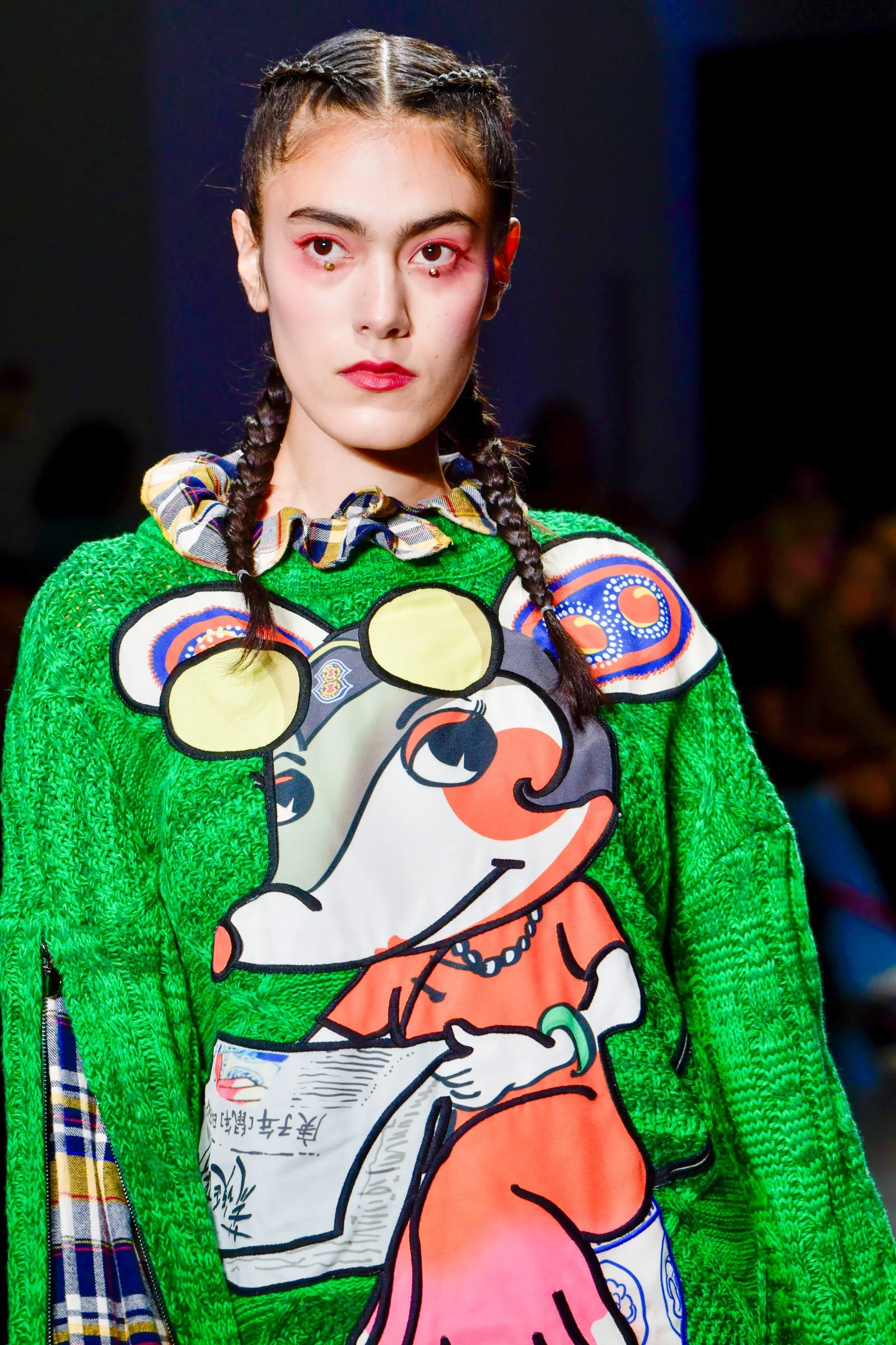 A young woman with brown braided hair and red makeup on her eyes and lips, wearing a bright green textured sweater with a graphic cartoon character on it, and a plaid ruffled collar.