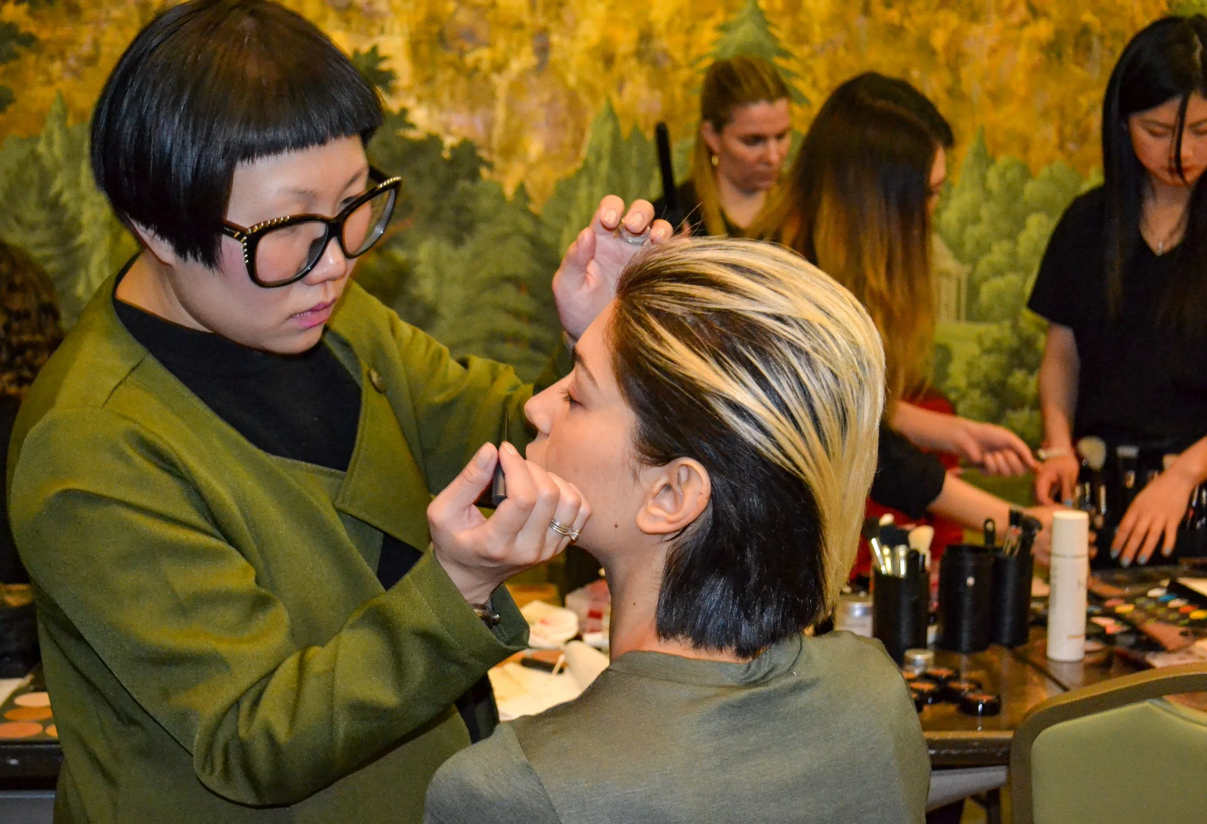 A woman with short black hair and glasses is applying makeup to another woman with short blond and black hair, seated at a makeup station. Several women are in the background preparing or applying makeup in a room with a nature-themed mural. 