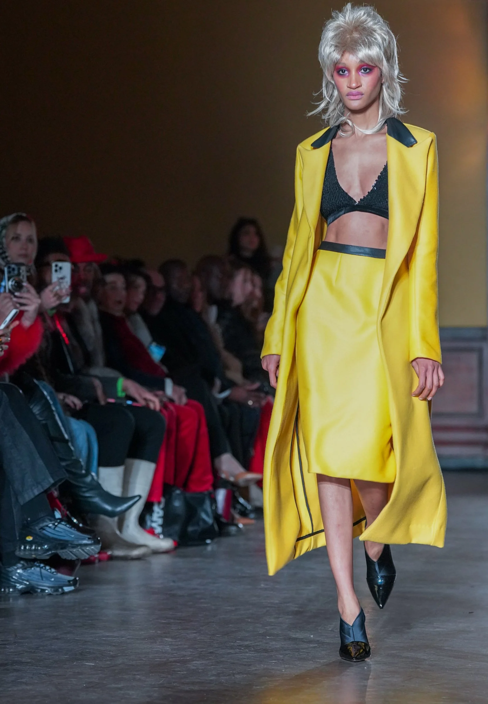 Model walking down runway in yellow coat with black accents, black bra top, black heels, and audience watching and taking photos.