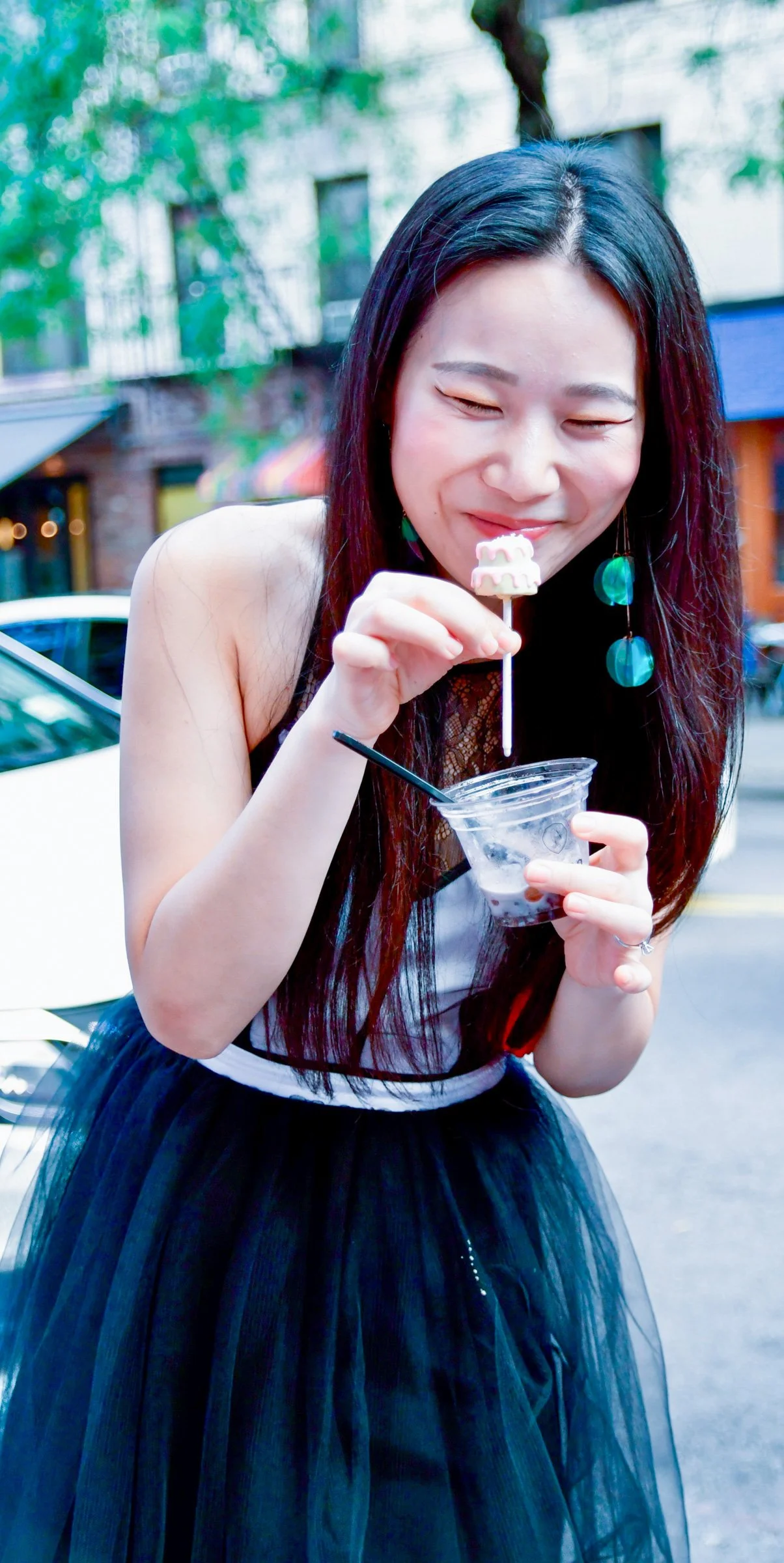 Young woman with long dark hair, wearing a black dress with a tulle skirt and turquoise earrings, smiling and holding a cup of ice cream with a small marshmallow or cookie on a stick outdoors. Grand Opening of Bar Pa Tea shop in Nolita, NYC.