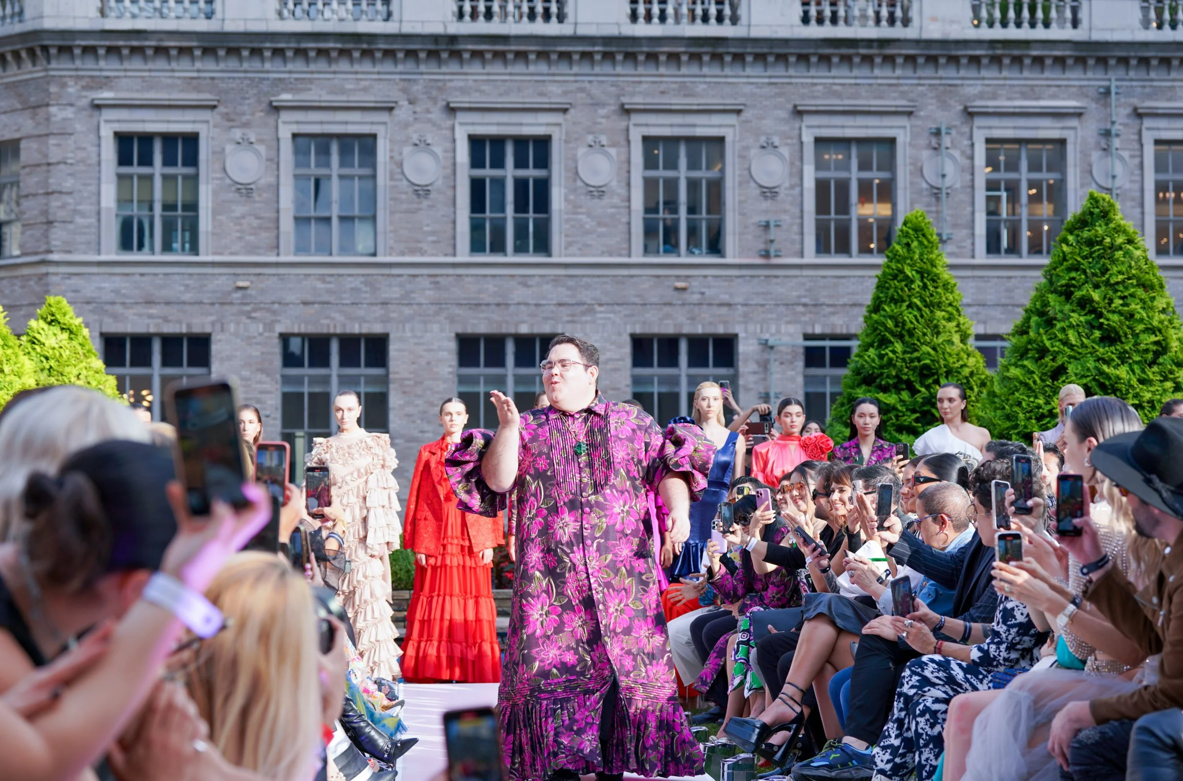 A man (Carlos Pineda) in a vibrant purple and pink patterned dress performing for a crowd at an outdoor event in front of a large brick building.