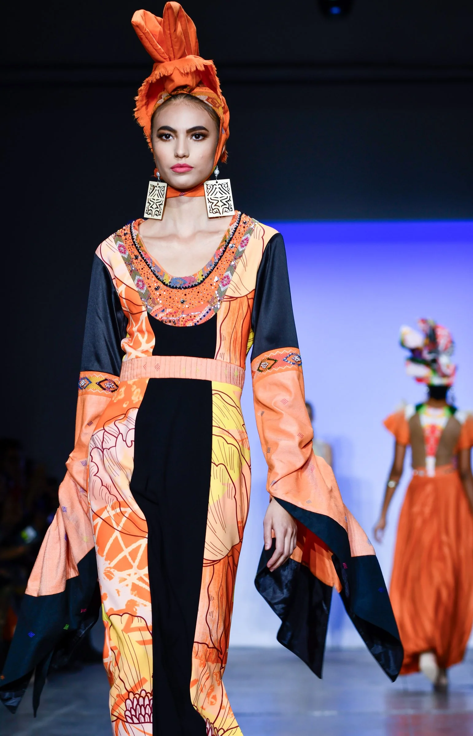 A female model walks down a runway wearing a colorful, patterned dress with orange, black, and yellow designs. She is accessorized with large rectangular earrings and an orange headwrap tied in a bow and styled as a turban. The background shows other