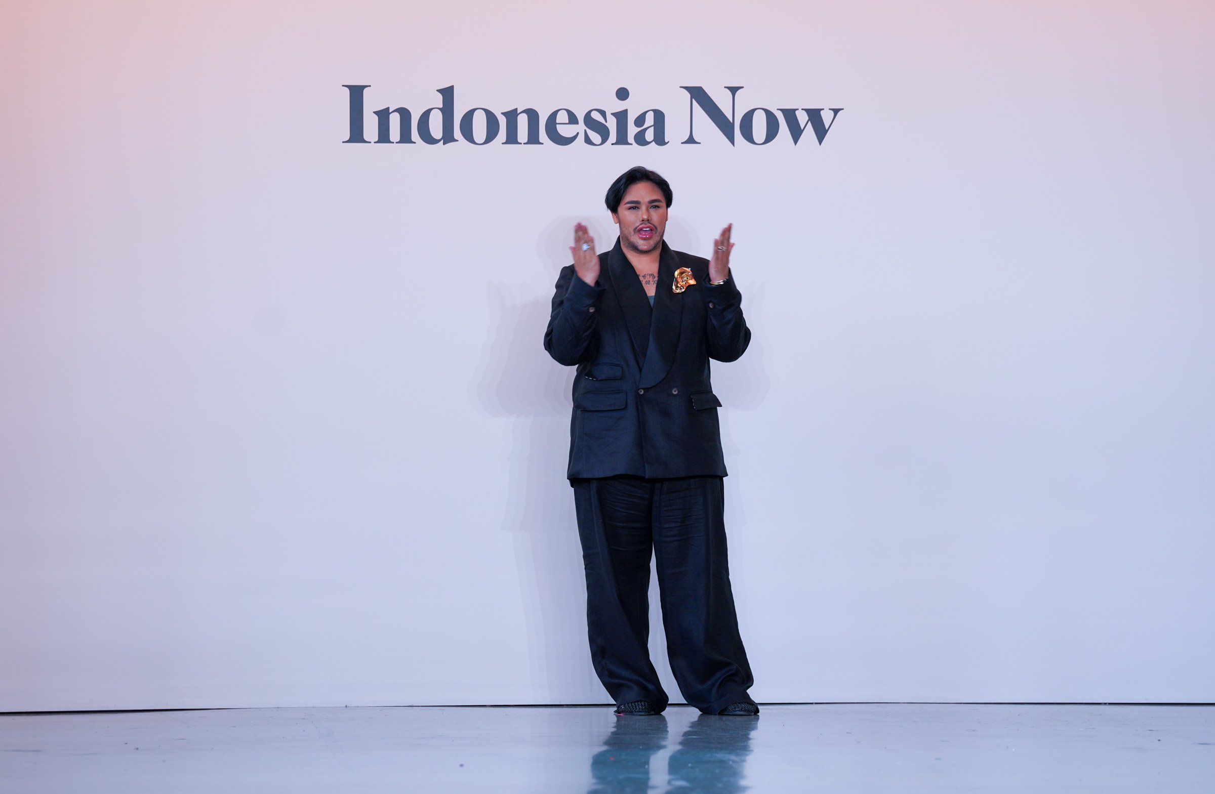 A person with dark hair and a beard standing in front of a white wall that reads 'Indonesia Now'. The person is wearing a black suit with a decorative gold pin or brooch on the lapel and appears to be speaking or gesturing with their hands.