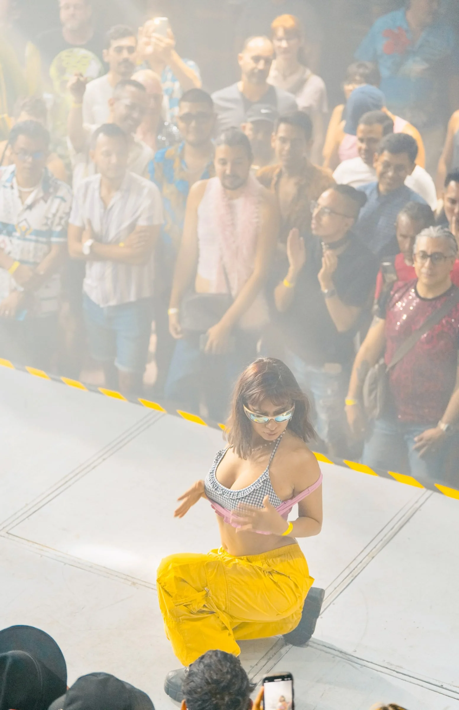 A young woman with sunglasses, a checkered crop top, and yellow baggy pants dancing in front of a reflective glass wall with a crowd of people behind her, some taking photos.