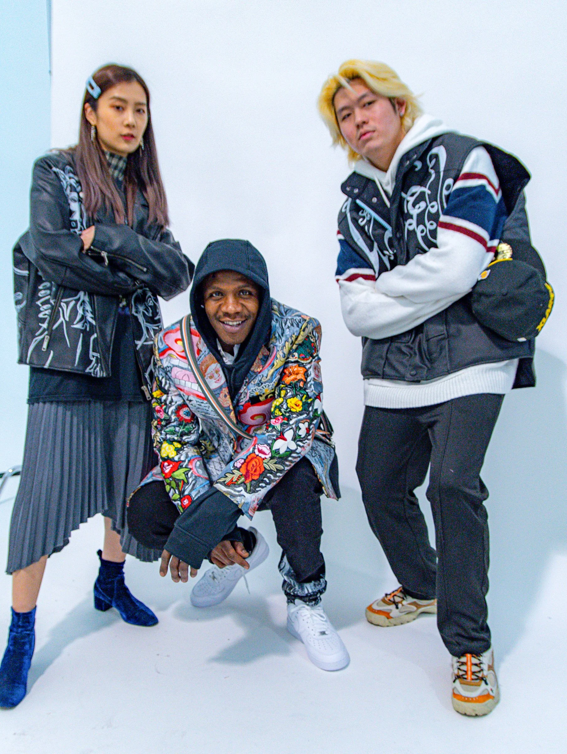 Four young people, three men and one woman, posing on a plain white background. They are dressed in casual streetwear with colorful and patterned clothing. The man on the right has blonde hair and is wearing a black vest with embroidery, gray pants, 