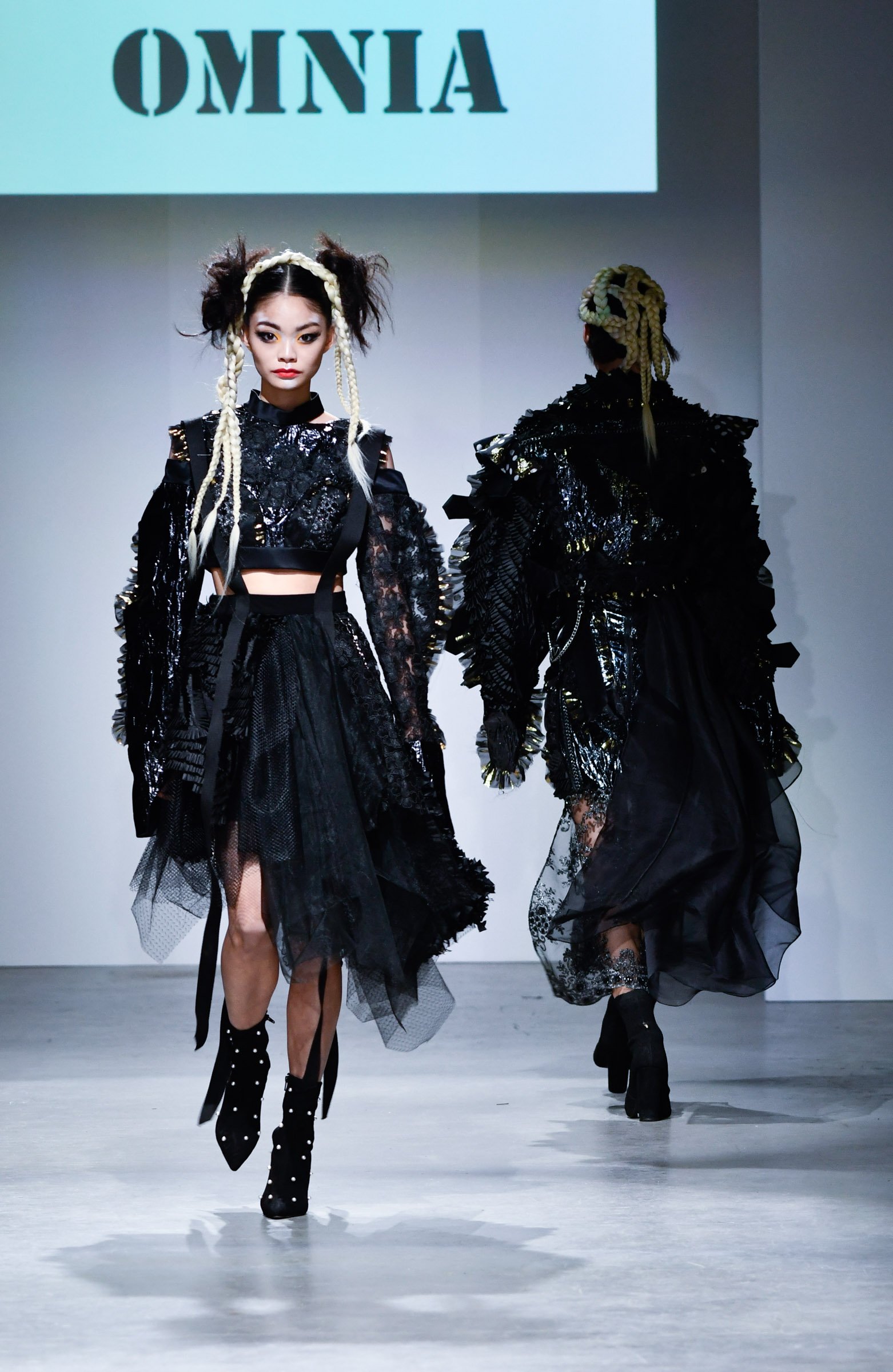 Two models walk down a runway during a fashion show. Both are wearing black, elaborate, textured outfits with sheer and lace details, paired with tall black heeled boots. The model in front has styled her hair in voluminous pigtails with blonde braid